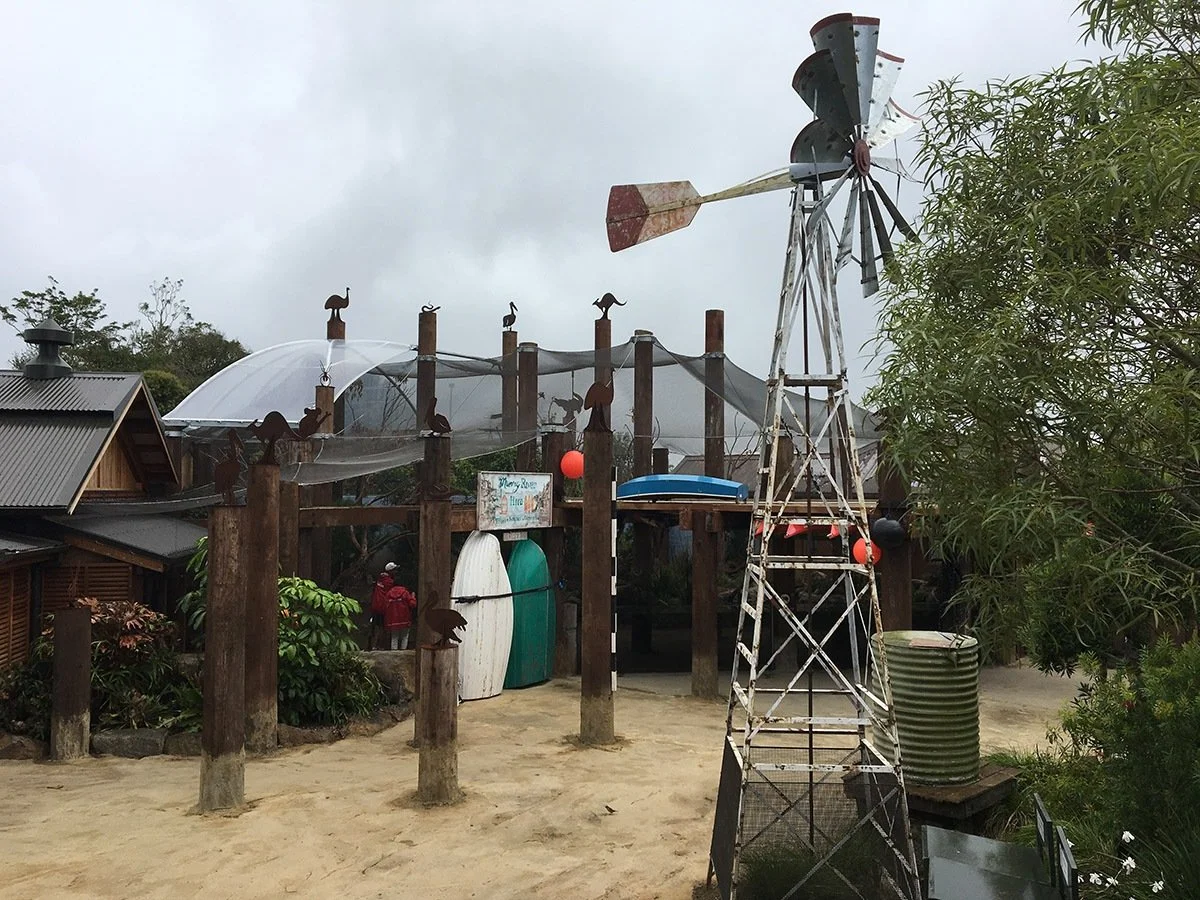 Outdoor playground with a wooden fence decorated with animal silhouettes, a windmill, surfboards, and lush greenery under a cloudy sky.