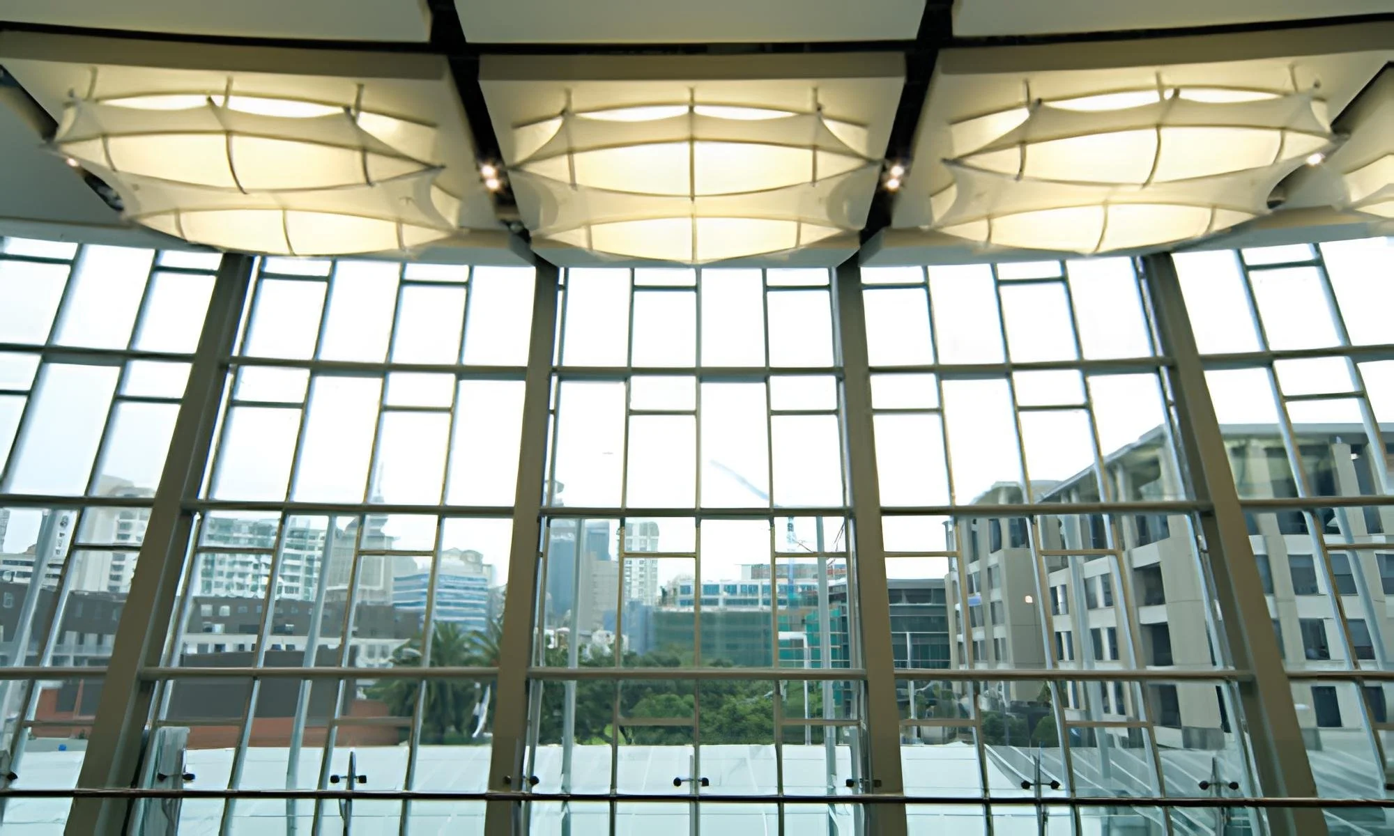 Interior of a modern building with large floor-to-ceiling windows and ceiling lights, overlooking an urban cityscape with high-rise buildings and greenery.