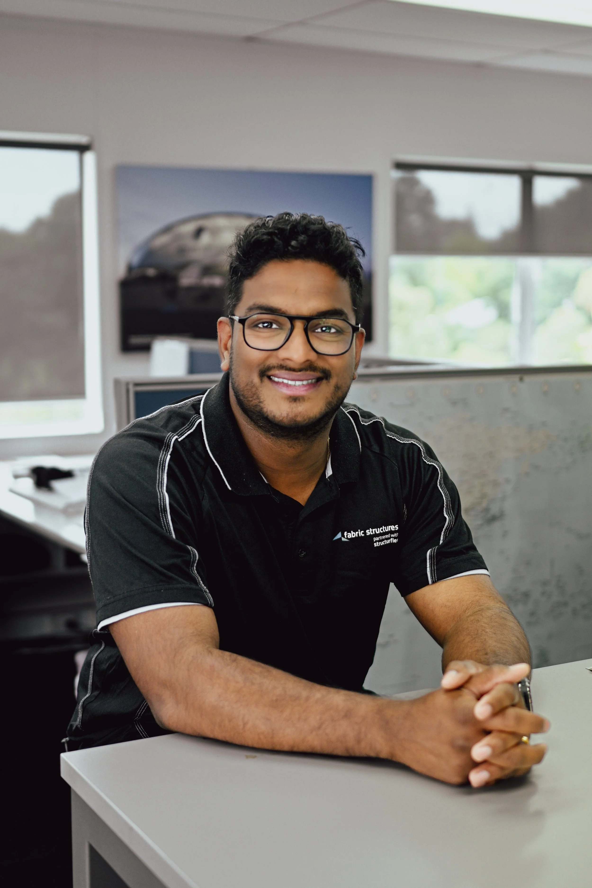 Nikhil Shine, Site Manager. A smiling man wearing glasses and a black polo shirt sitting at a desk in an office environment.