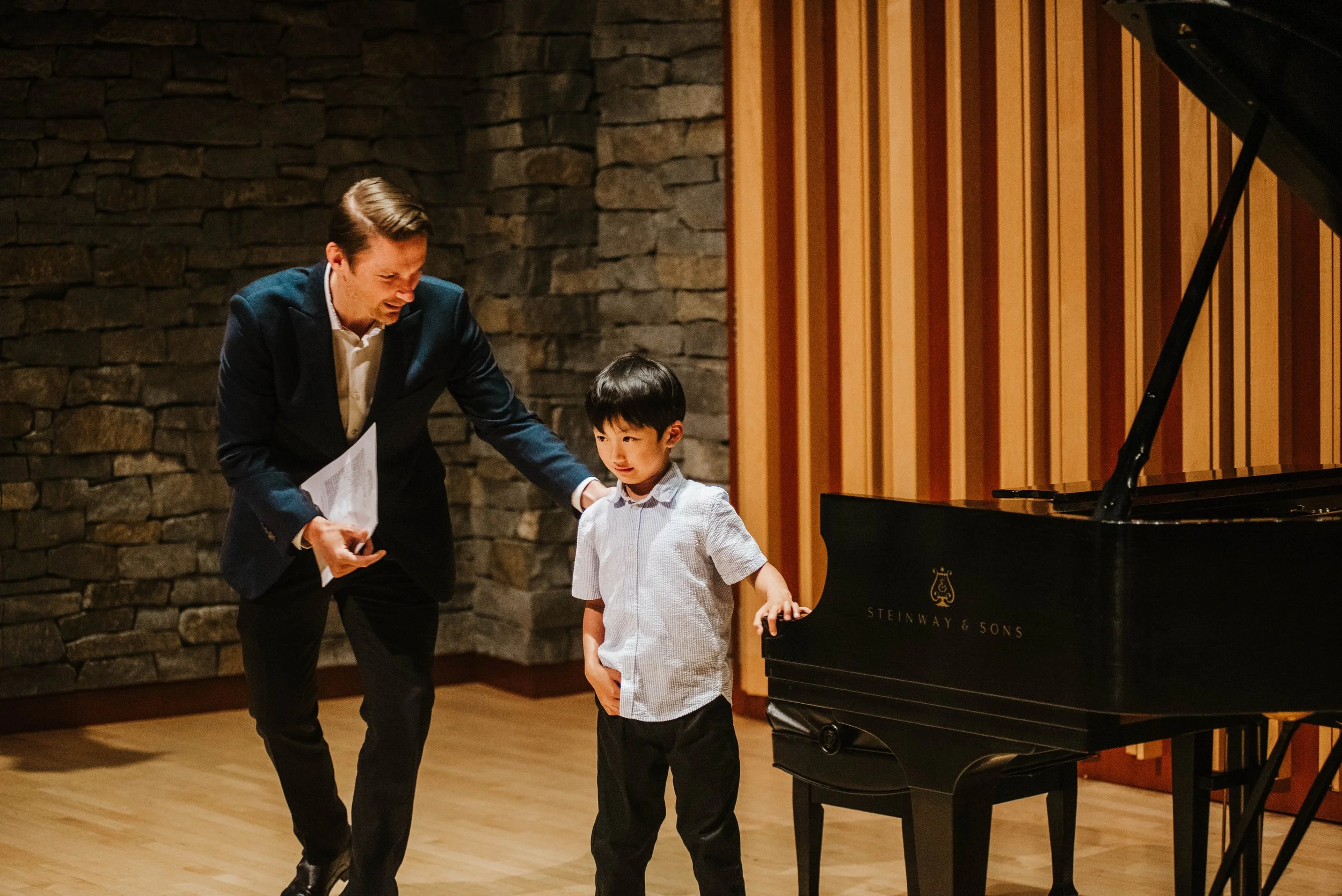 A man dressed in a dark suit coaching a young boy at a grand piano in a music studio with a stone wall and wooden wall paneling.