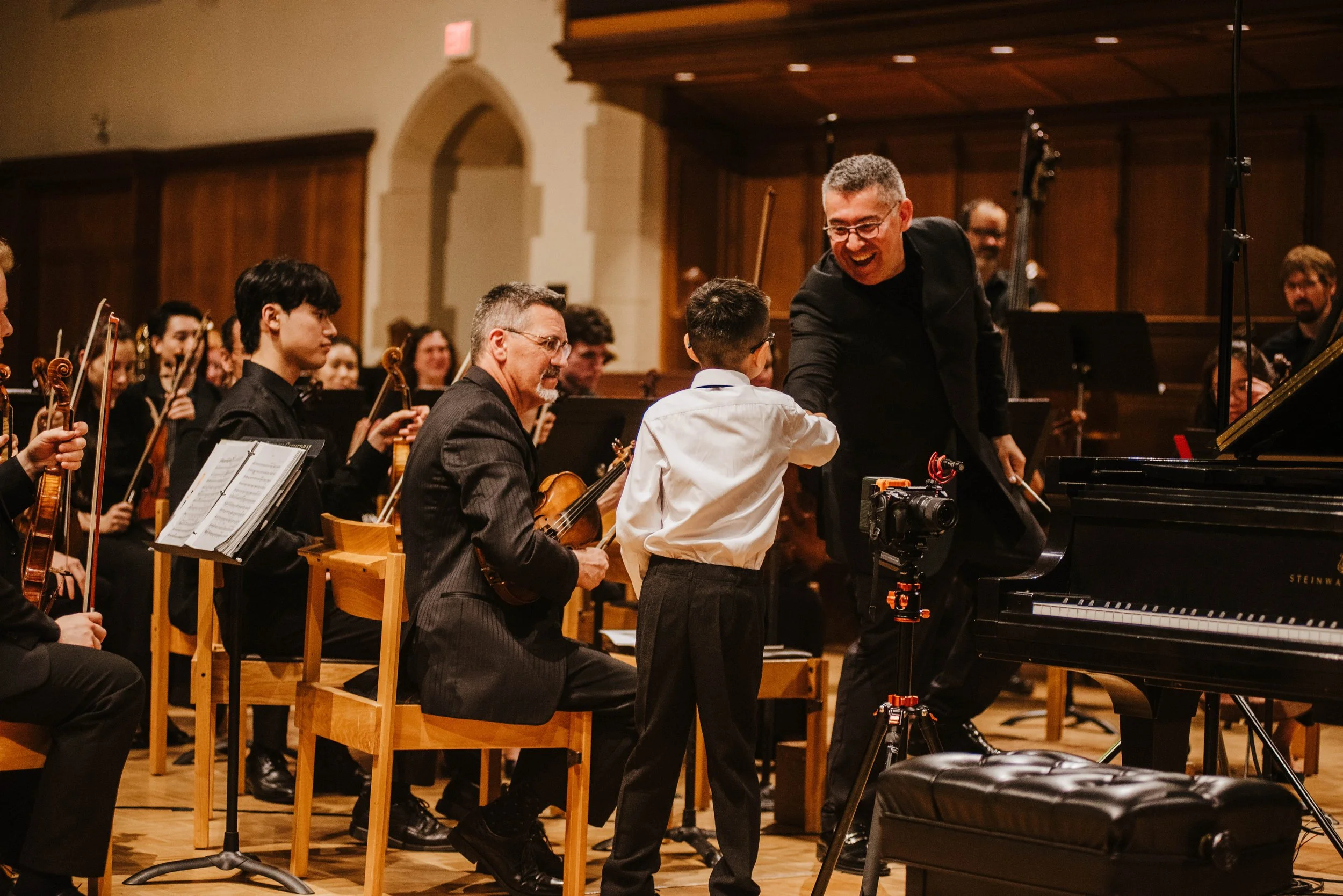 A young boy in a white shirt shaking hands with a man in a black jacket next to a piano during an orchestral concert.