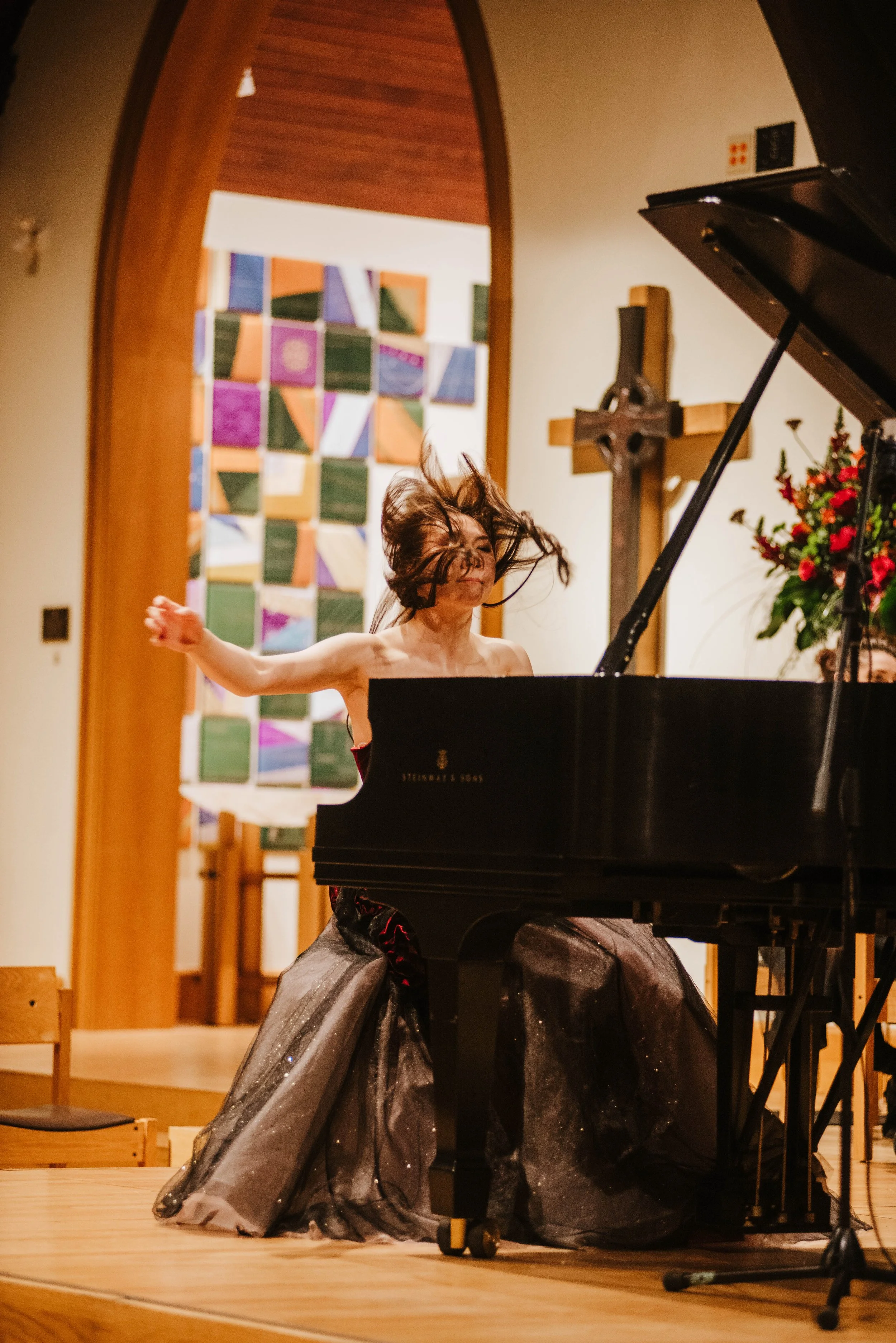 A woman in a black dress is playing a grand piano on stage, her hair flying as she passionately performs.
