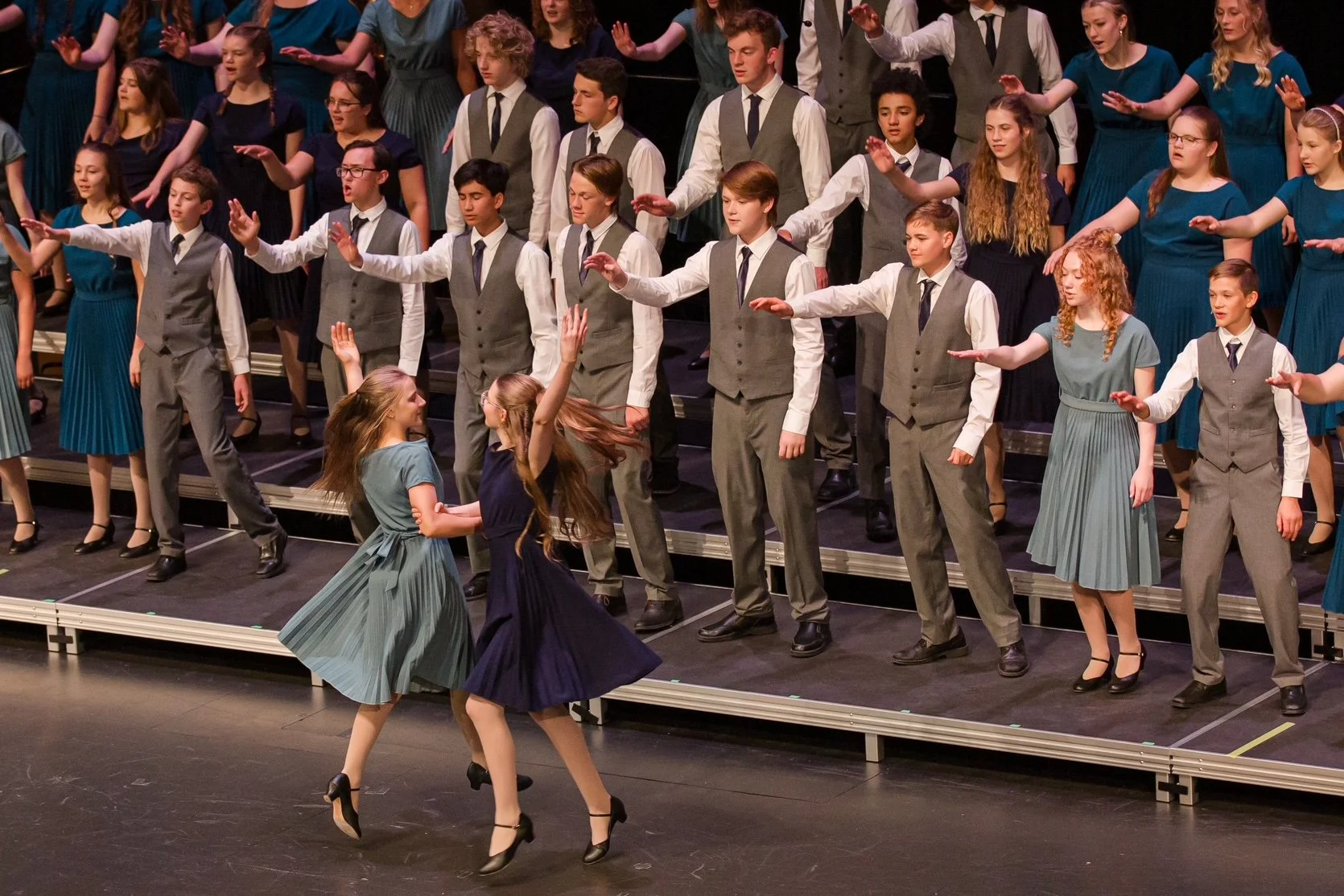 Children in formal attire performing a choreographed dance on a theater stage during a school or community concert.
