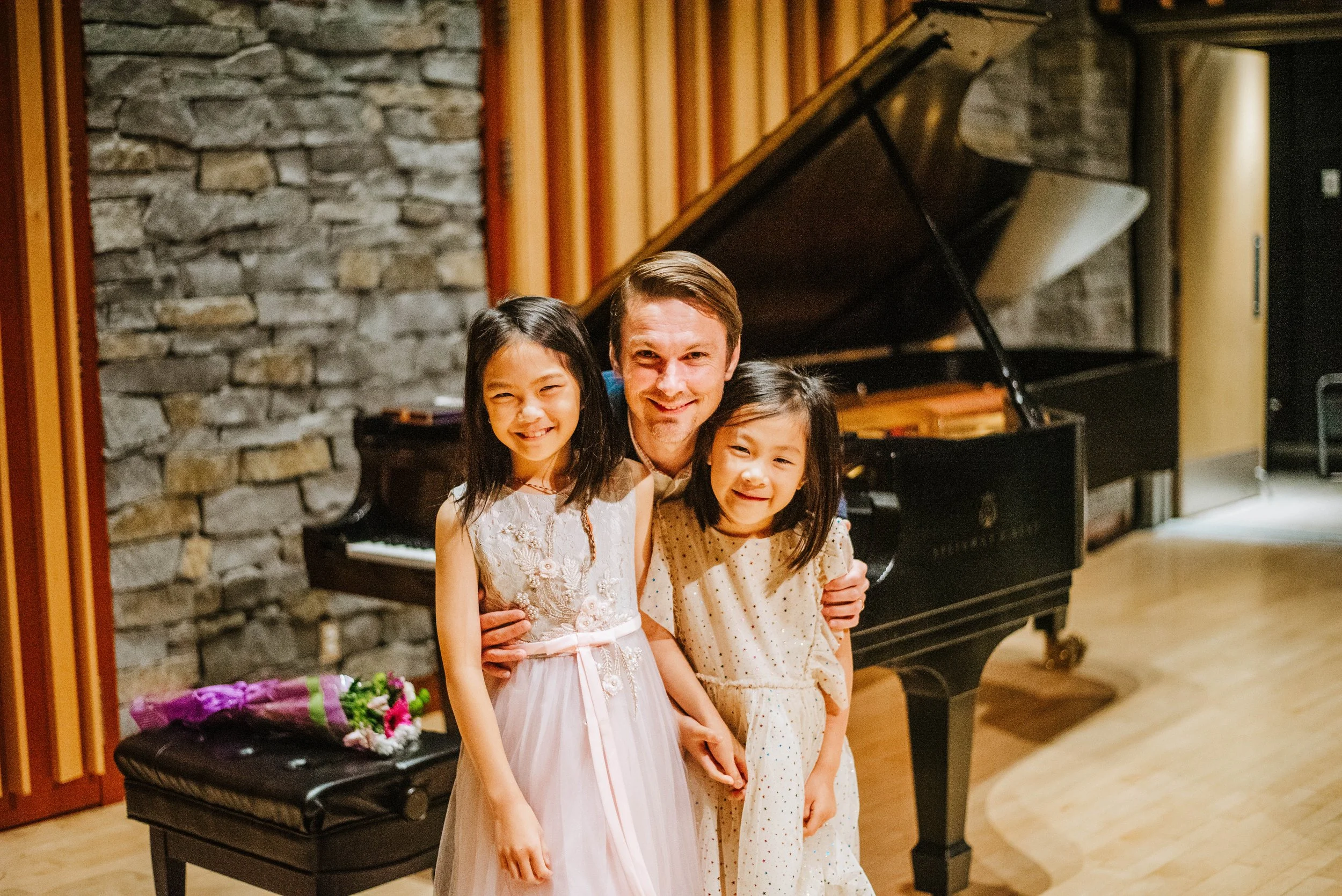 A man with two young girls smiling and posing in front of a grand piano in a cozy room with stone and wood walls.
