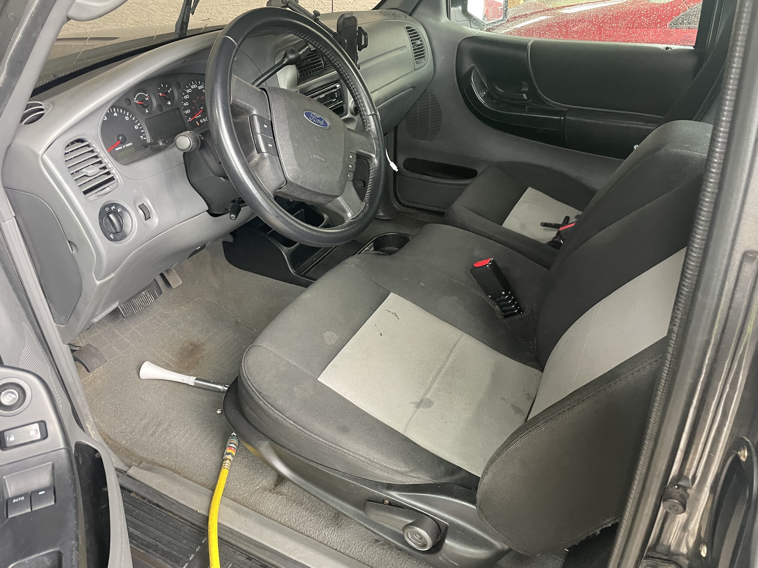 Interior of a vehicle showing driver's seat, steering wheel, dashboard, and a hose on the floor.