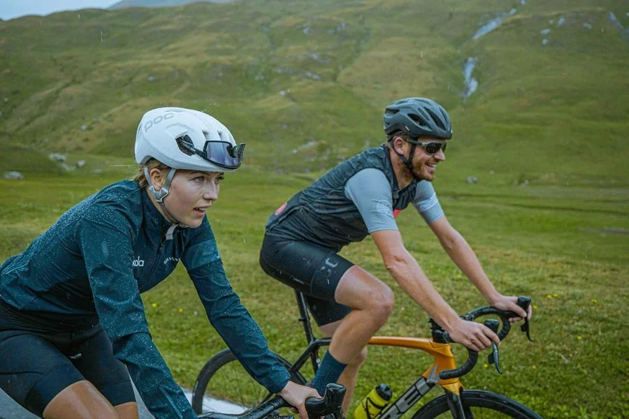Two cyclists ride through a grassy hillside landscape with green hills and small streams in the background, wearing helmets and cycling gear.