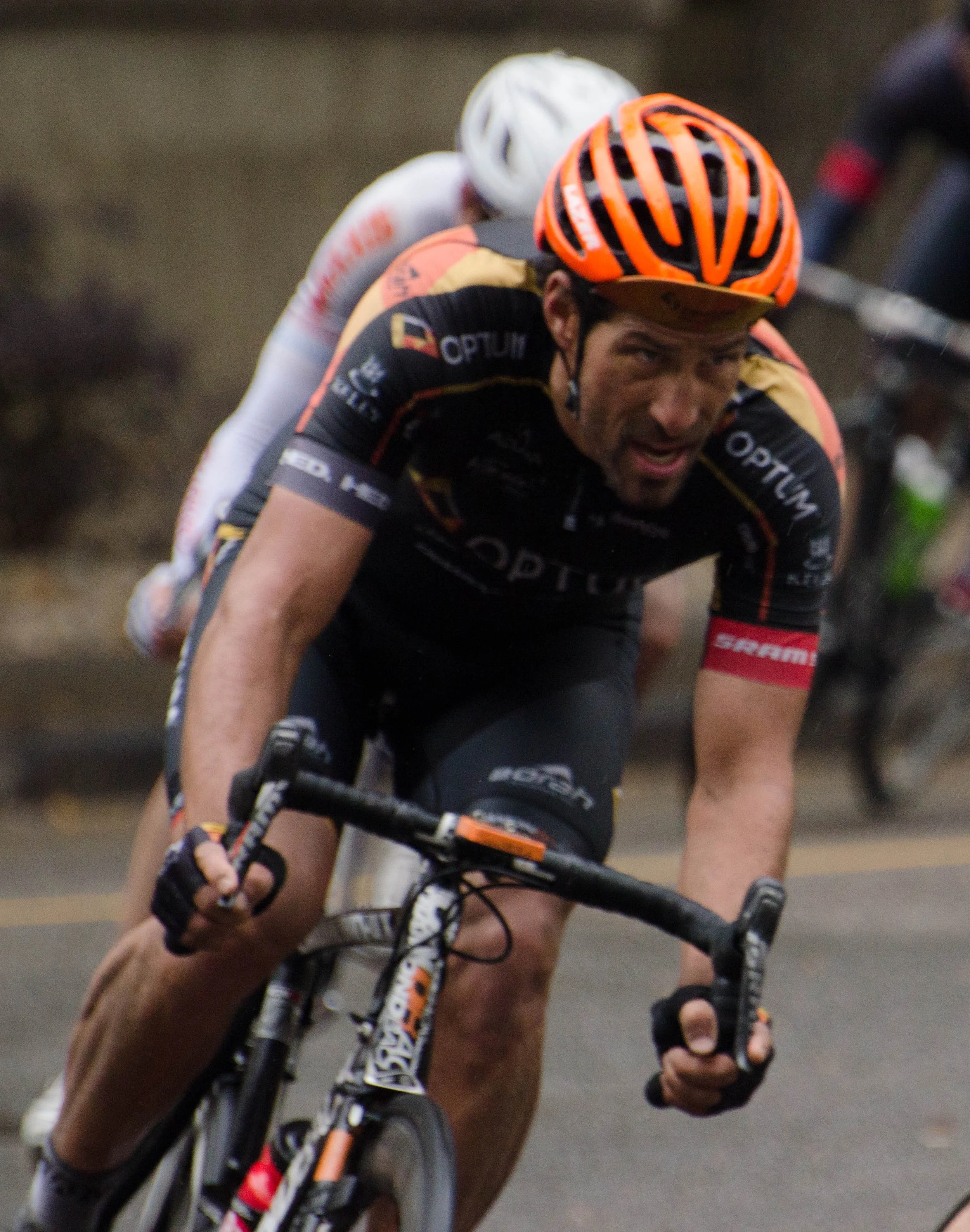 A male cyclist wearing a black Optum team jersey, orange helmet, and black biking gloves, intensely focused during a race, with other cyclists visible in the background.