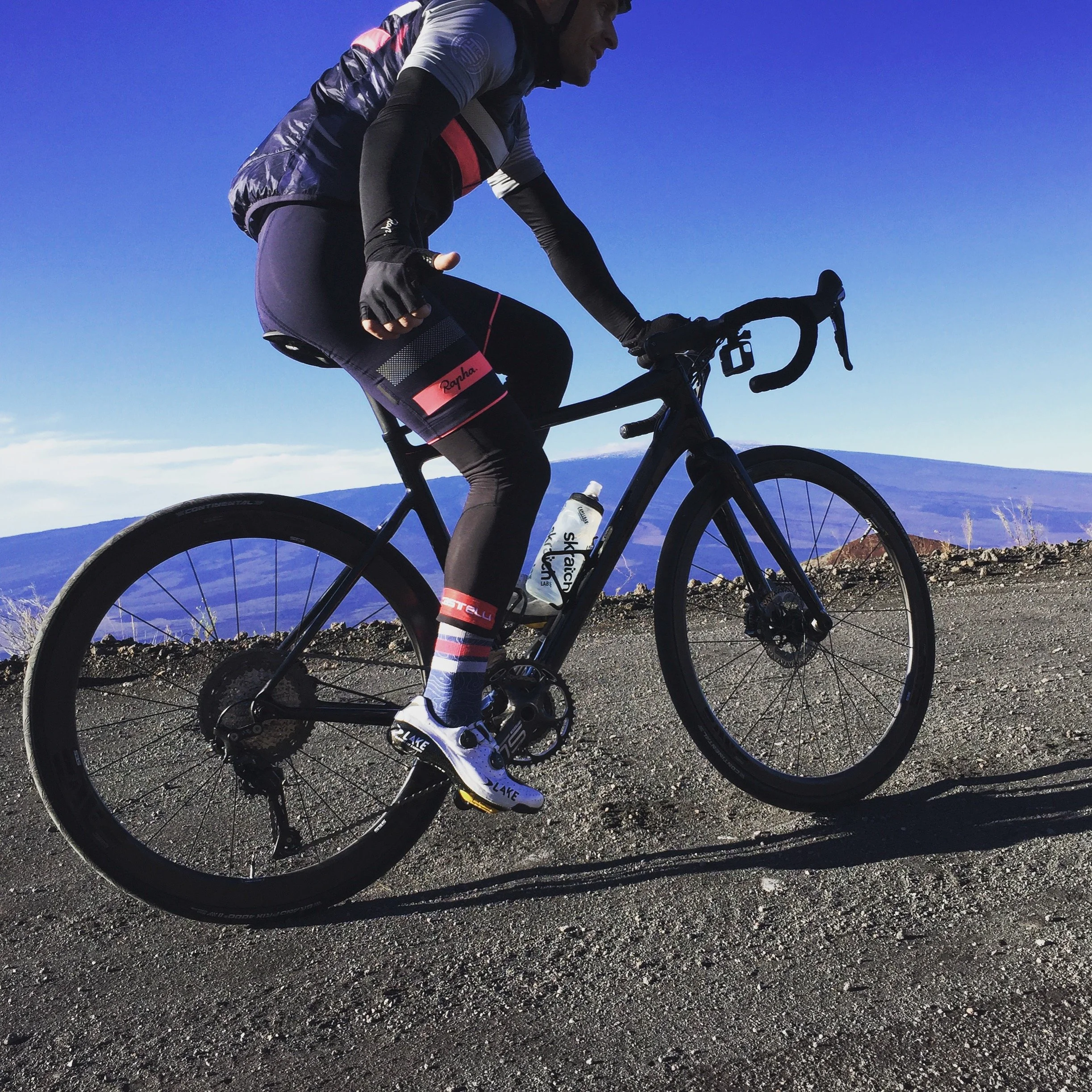 A cyclist riding a black bicycle on a dirt trail with a mountain and clear blue sky in the background.