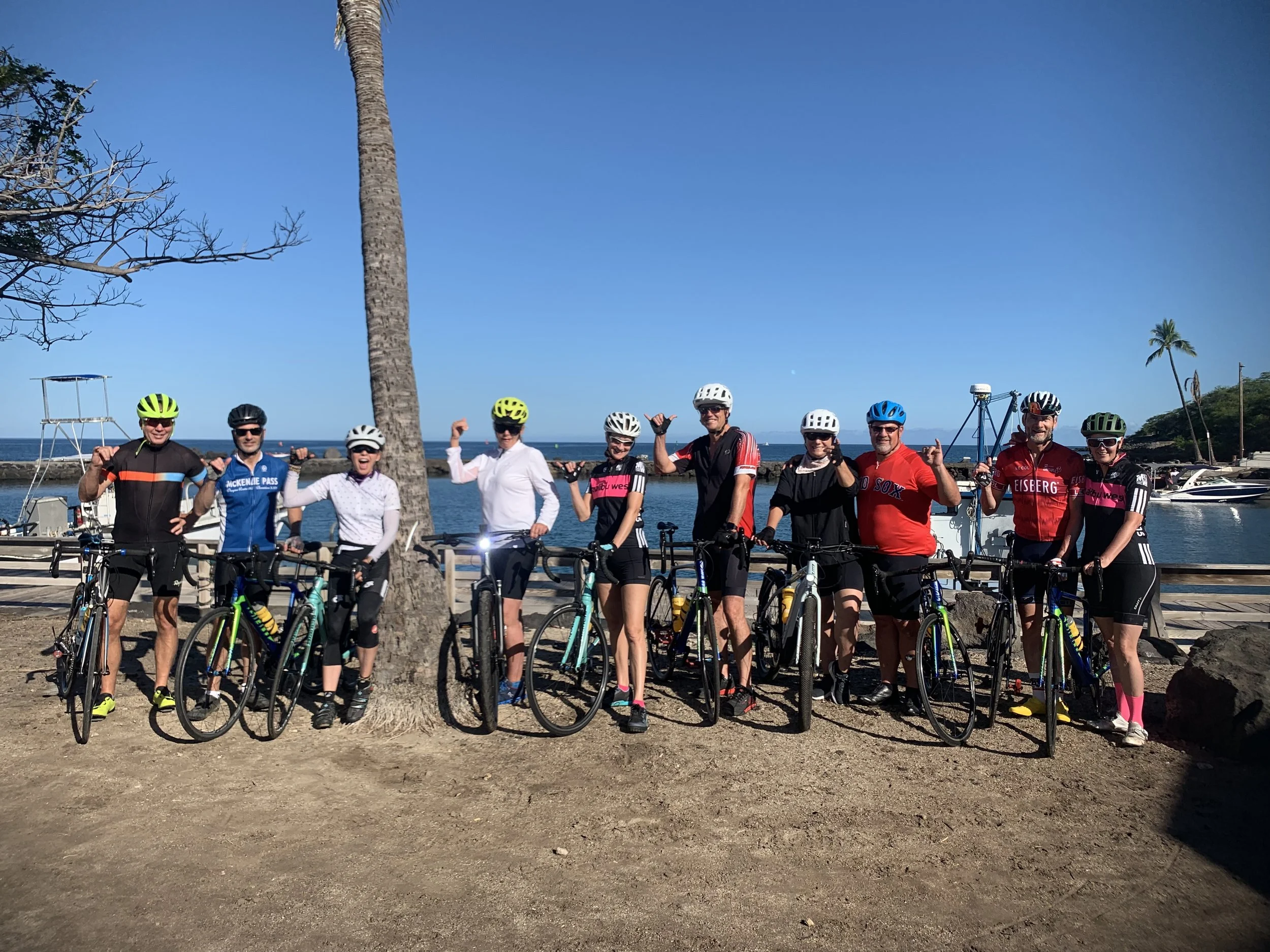 Group of ten cyclists in cycling gear and helmets standing with bikes near a palm tree by a marina with boats, on a sunny day with clear blue sky.
