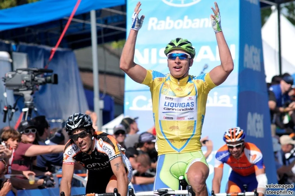 Cyclist in a yellow and blue jersey raising his arms in victory at a race, with other cyclists and spectators in the background.