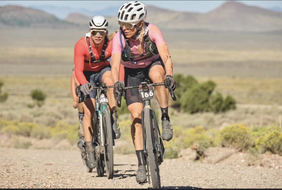 Two women mountain biking on a dirt trail in a desert landscape, wearing helmets and sports gear.