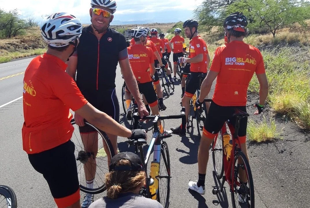 A group of cyclists wearing red jerseys with yellow text, gathered on the side of a road with a scenic landscape, some wearing helmets and sunglasses, engaged in conversation, with one cyclist in a black jersey.