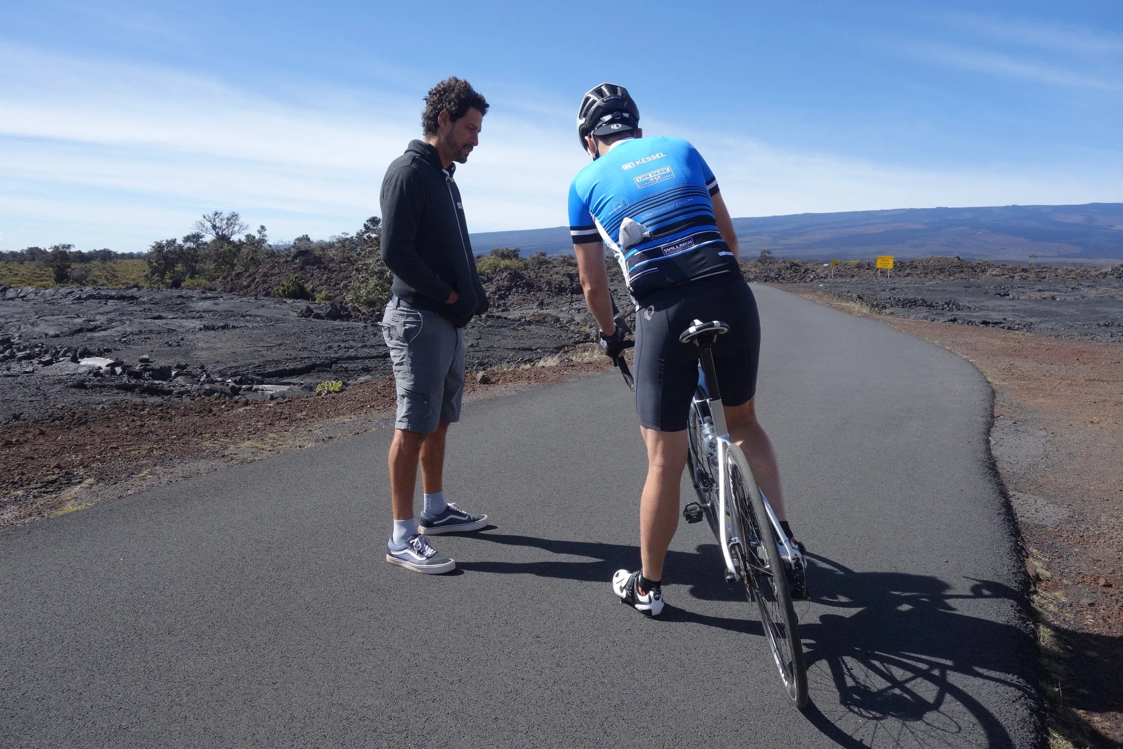 A person in cycling gear, wearing a helmet, stands next to a bicycle on a paved road in a volcanic landscape. Another man in casual clothing watches.