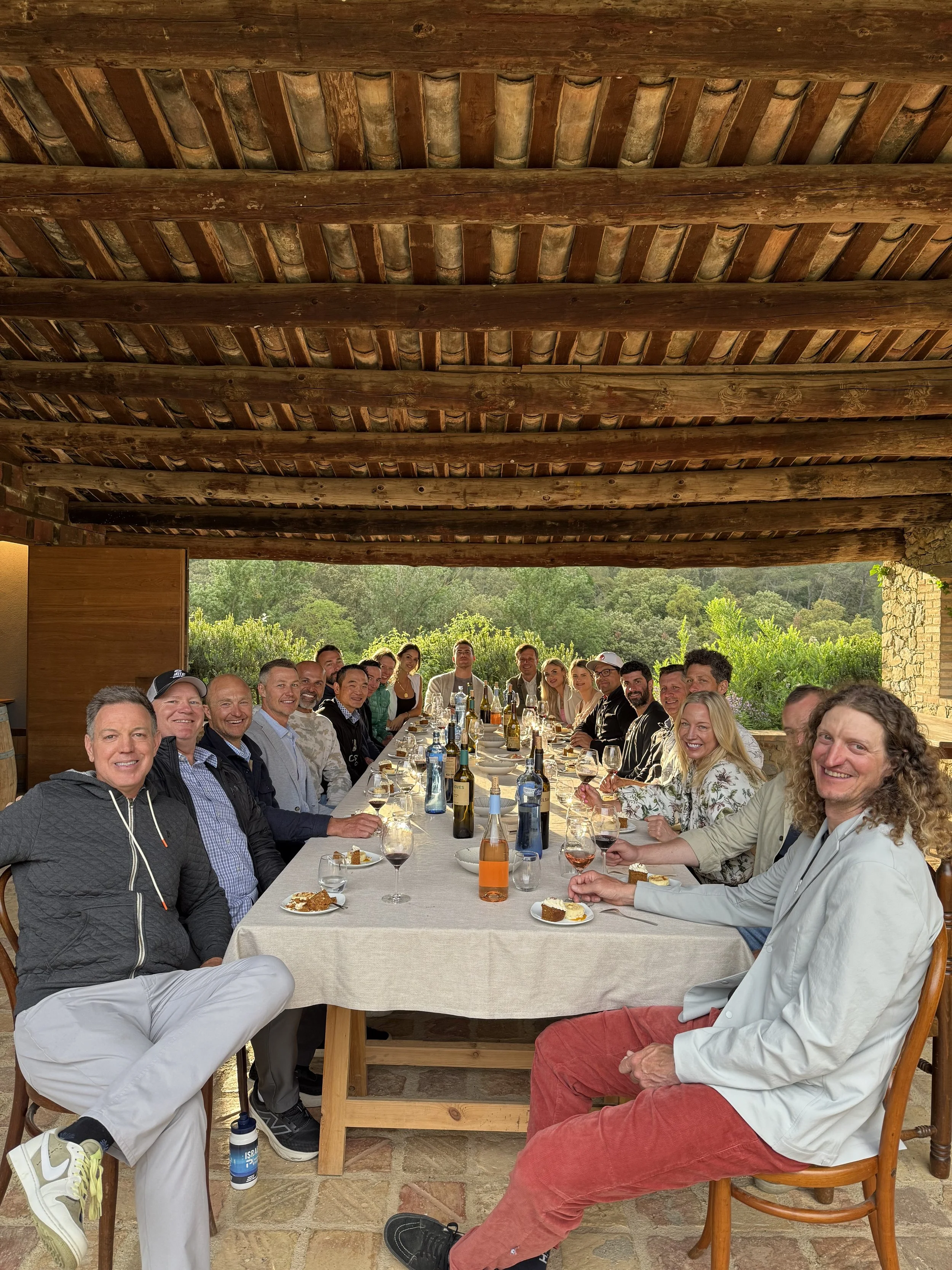 A large group of people gathered around a long dining table outdoors, enjoying a meal together. The setting is covered by a rustic wooden roof, with lush green trees and landscape in the background. The table is decorated with various bottles of wine and drinks, glasses, plates of food, and desserts. Everyone is smiling and appears to be having a good time.