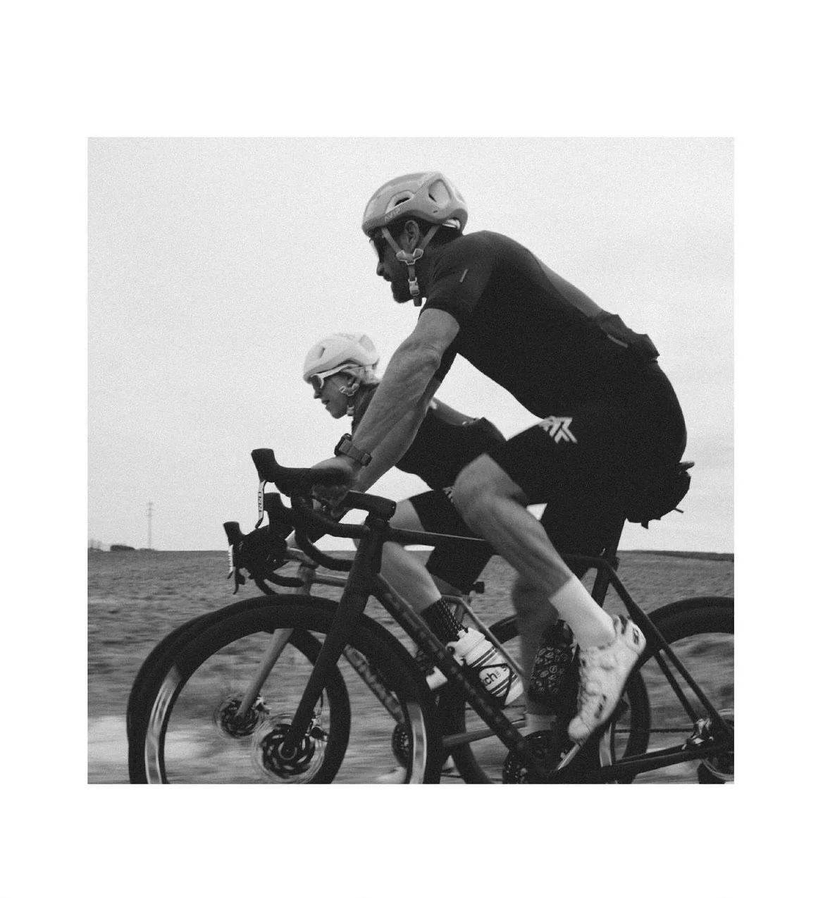 Two male cyclists riding mountain bikes on a trail, wearing helmets and athletic gear, black and white photograph.