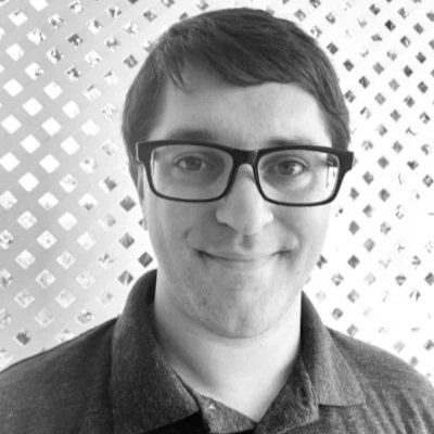 Black-and-white photo of a smiling man with short dark hair and glasses, wearing a dark collared shirt, standing in front of a patterned background.
