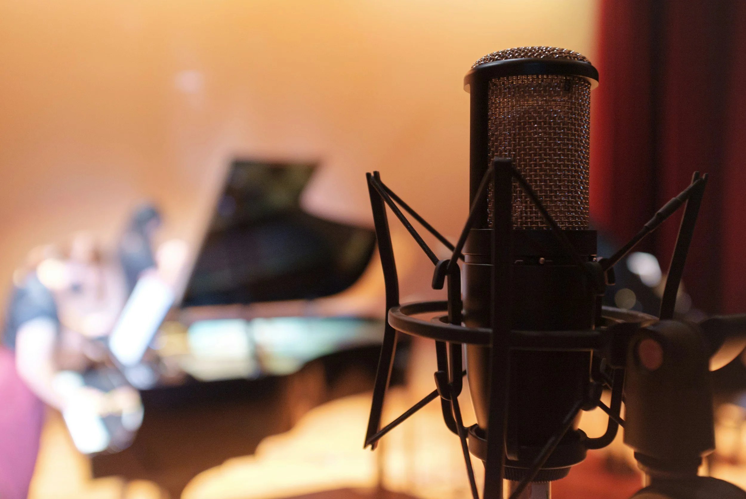 A close-up of a microphone in a recording studio with a blurred person playing a grand piano in the background
