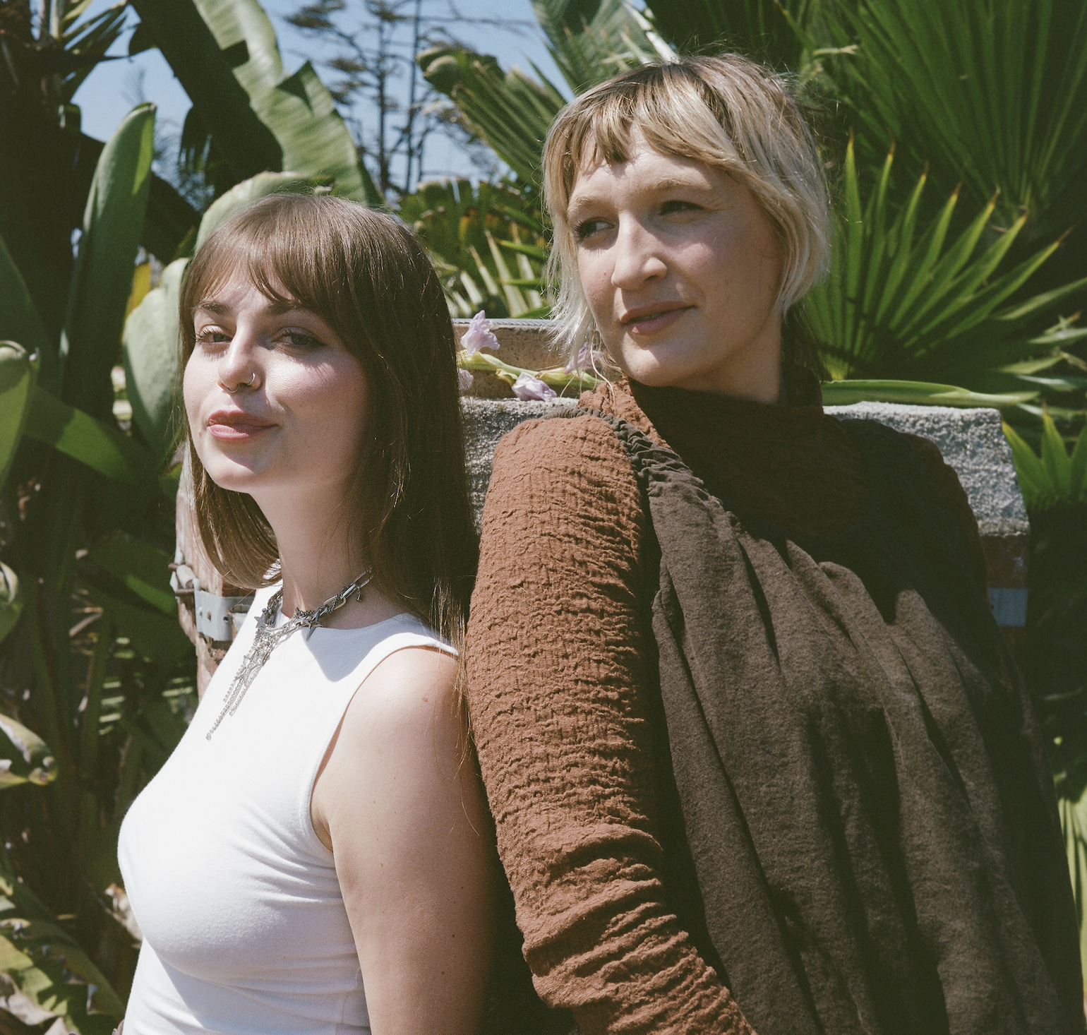 Two women standing side by side outdoors, with lush green tropical plants in the background, facing opposite directions but leaning back-to-back.