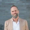 Smiling man with a beard wearing a blazer, standing against a brick wall.