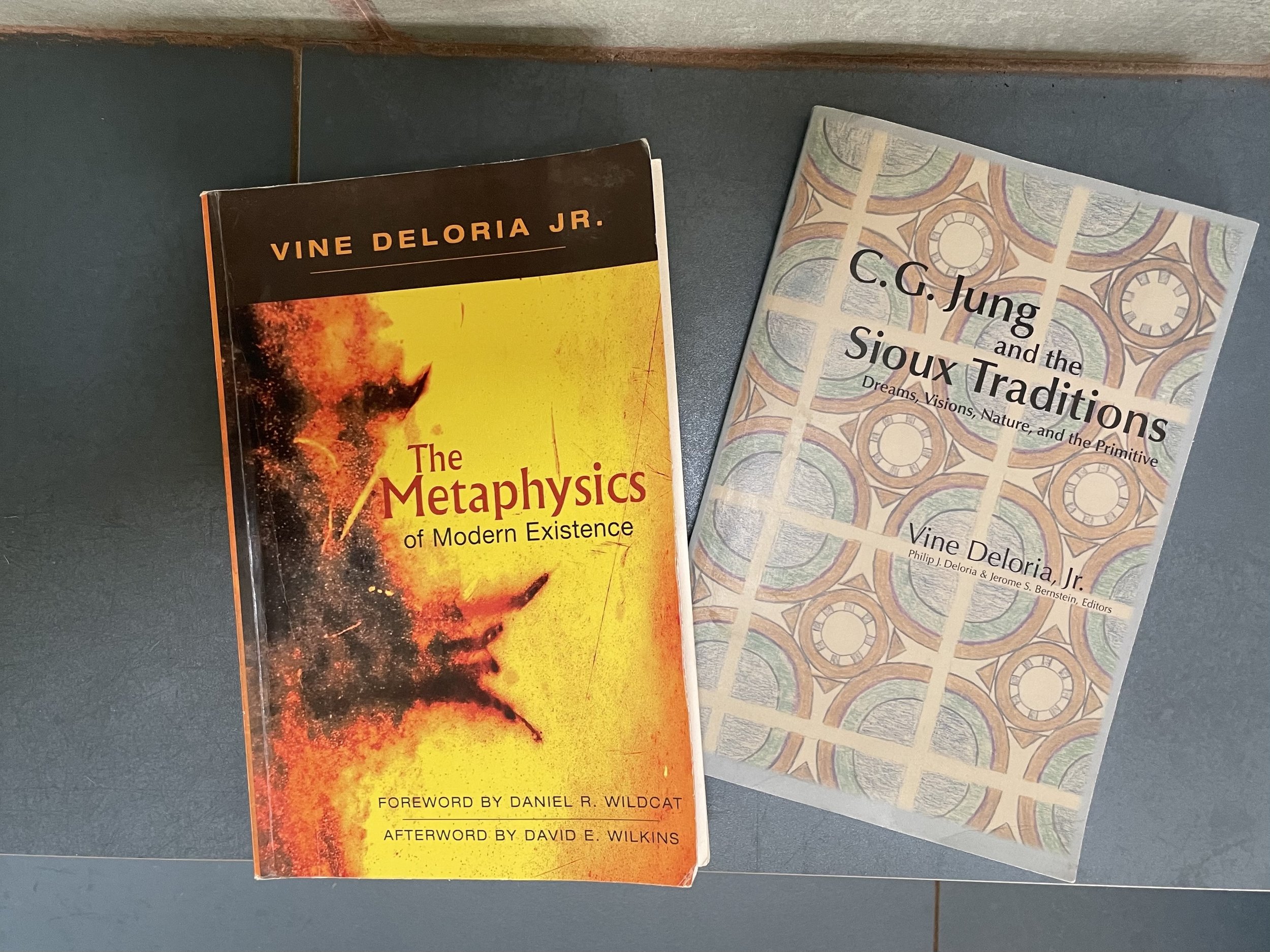 Two books on a dark surface. The left book is titled 'The Metaphysics' by Vine Deloria Jr. featuring a fiery illustration on the cover. The right book is titled 'C.C. Jung and the Sioux Traditions: Dreams, Visions, Nature, and the Primitive' with a patterned cover.