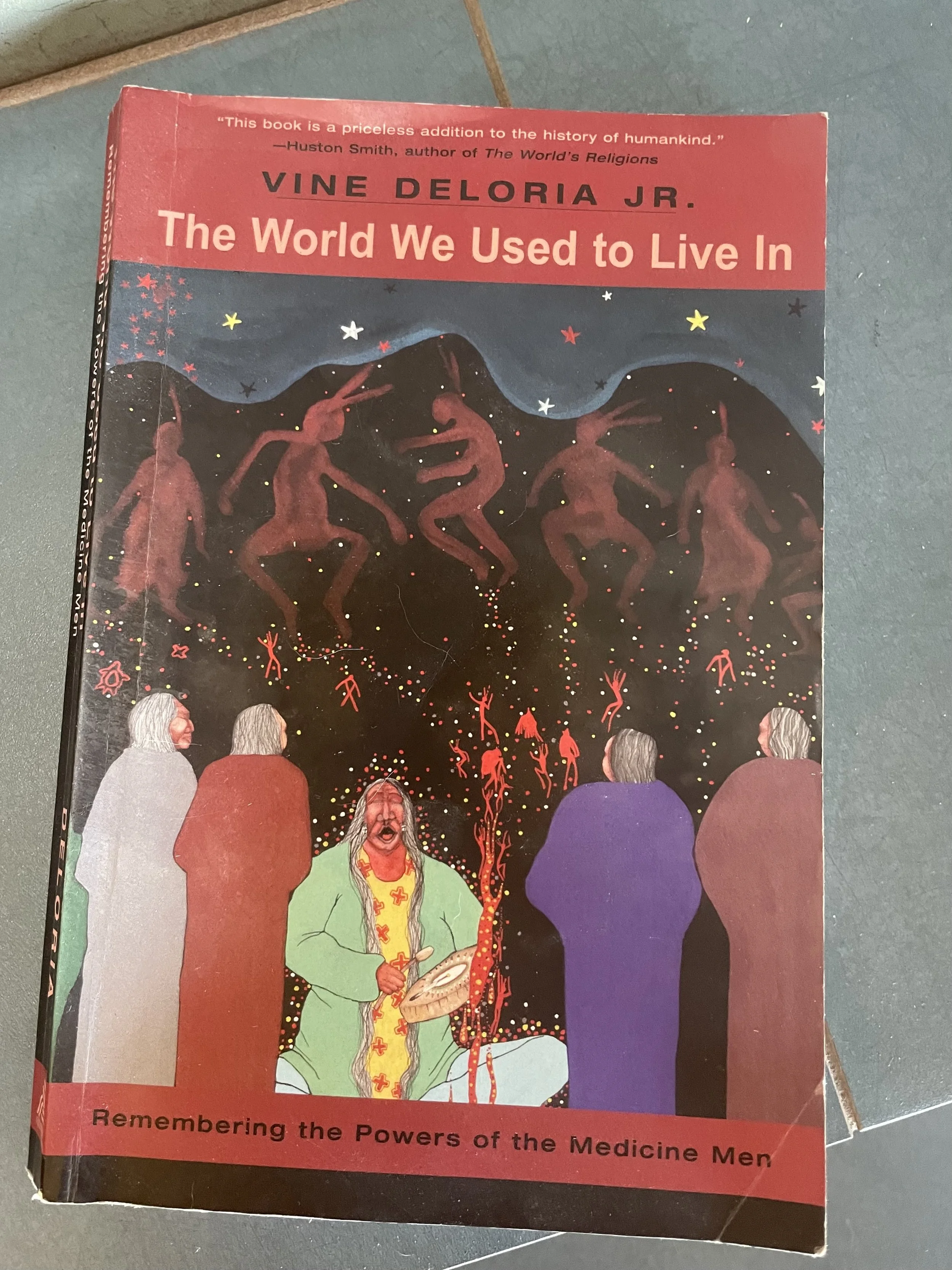 Book cover titled "The World We Used to Live In" by Vine Deloria Jr., featuring illustrations of Native American figures, a mountain with dancing figures, and a colorful, starry background.