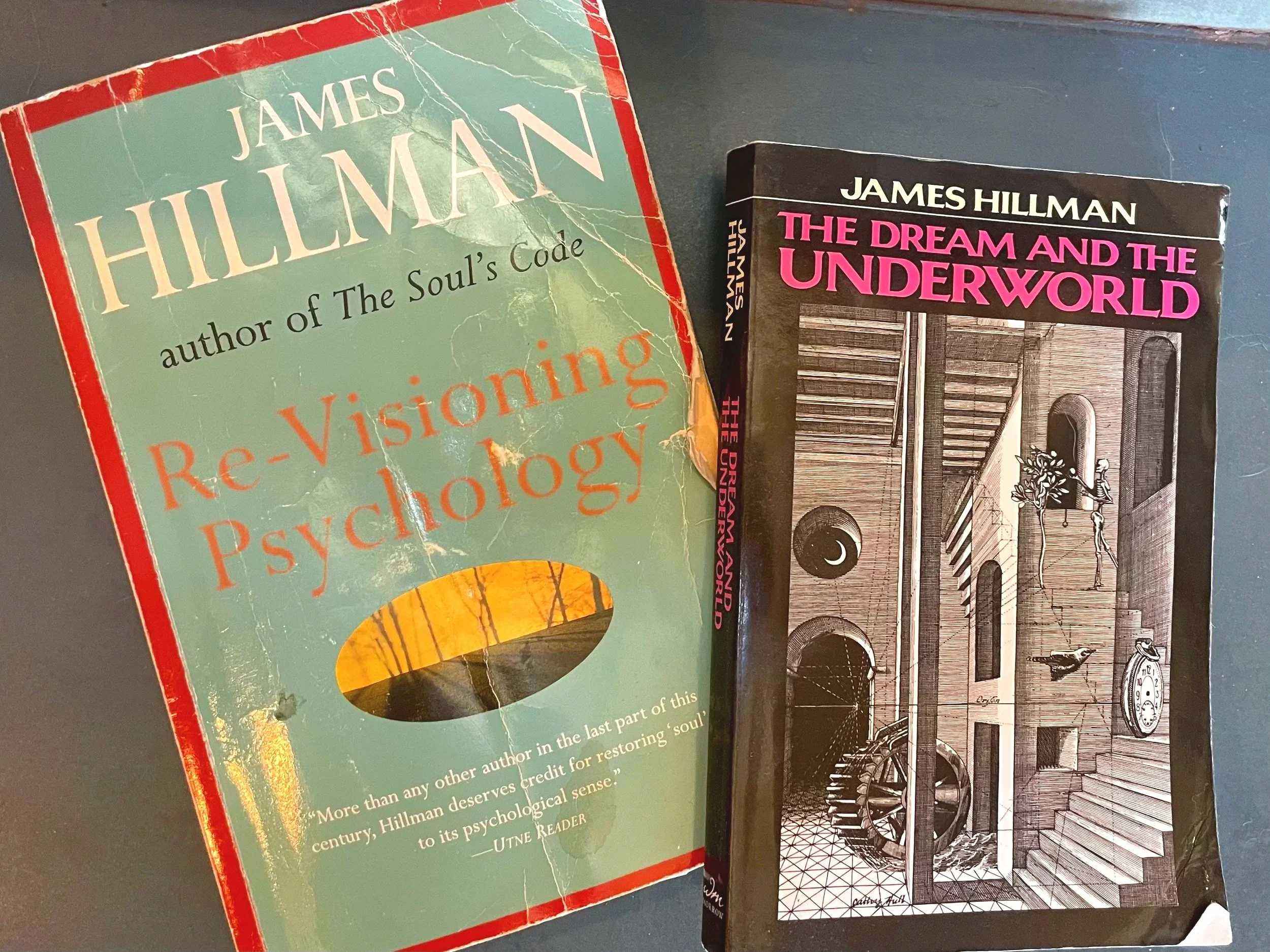 A book titled 'Re-Visions of Psychology' by James Hillman with a worn cover and visible creases, placed next to a black-and-white book titled 'The Dream and the Underworld' by James Hillman, featuring an illustration of an interior with spiral stairs, windows, and a person standing near a bicycle.
