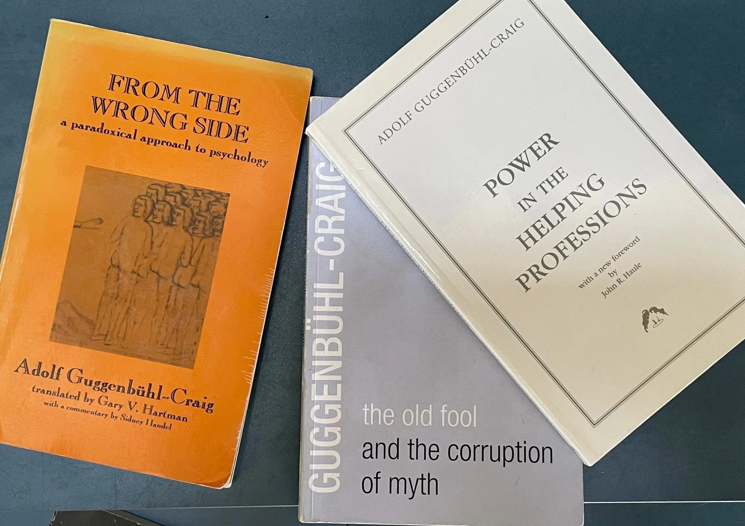 Three books on a dark blue surface. The left book is orange with black and red text, titled "From the Wrong Side: A Paradoxical Approach to Psychology" by Adolf Guggenbühl-Craig. The middle book is partly visible, gray with white text. The right book is white with black text, titled "Power in the Helping Professions" by Adolf Guggenbühl-Craig.