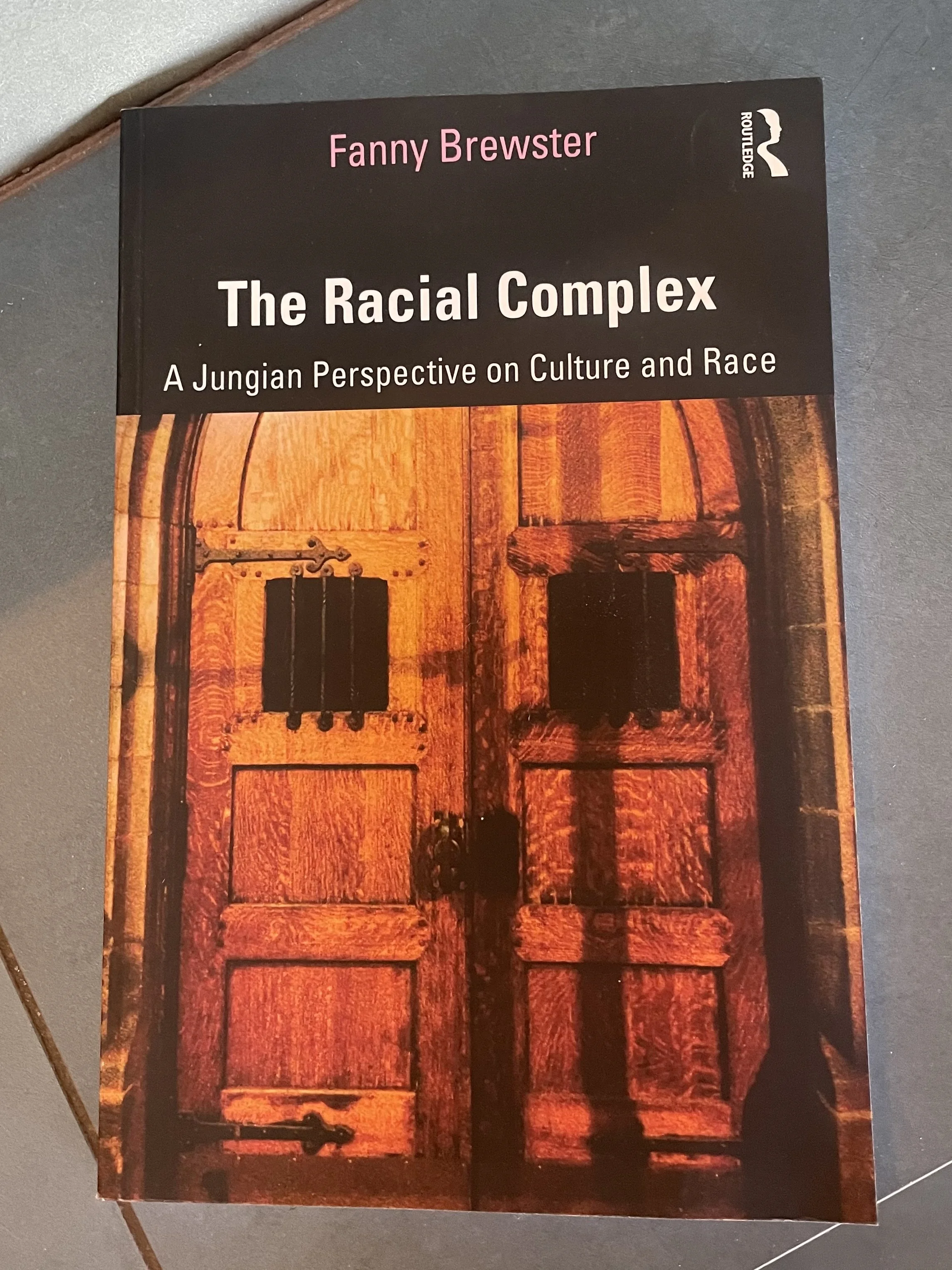 Book titled 'The Racial Complex: A Jungian Perspective on Culture and Race' by Fanny Brewster, published by Routledge, featuring a wooden door or window with black hinges on the cover.