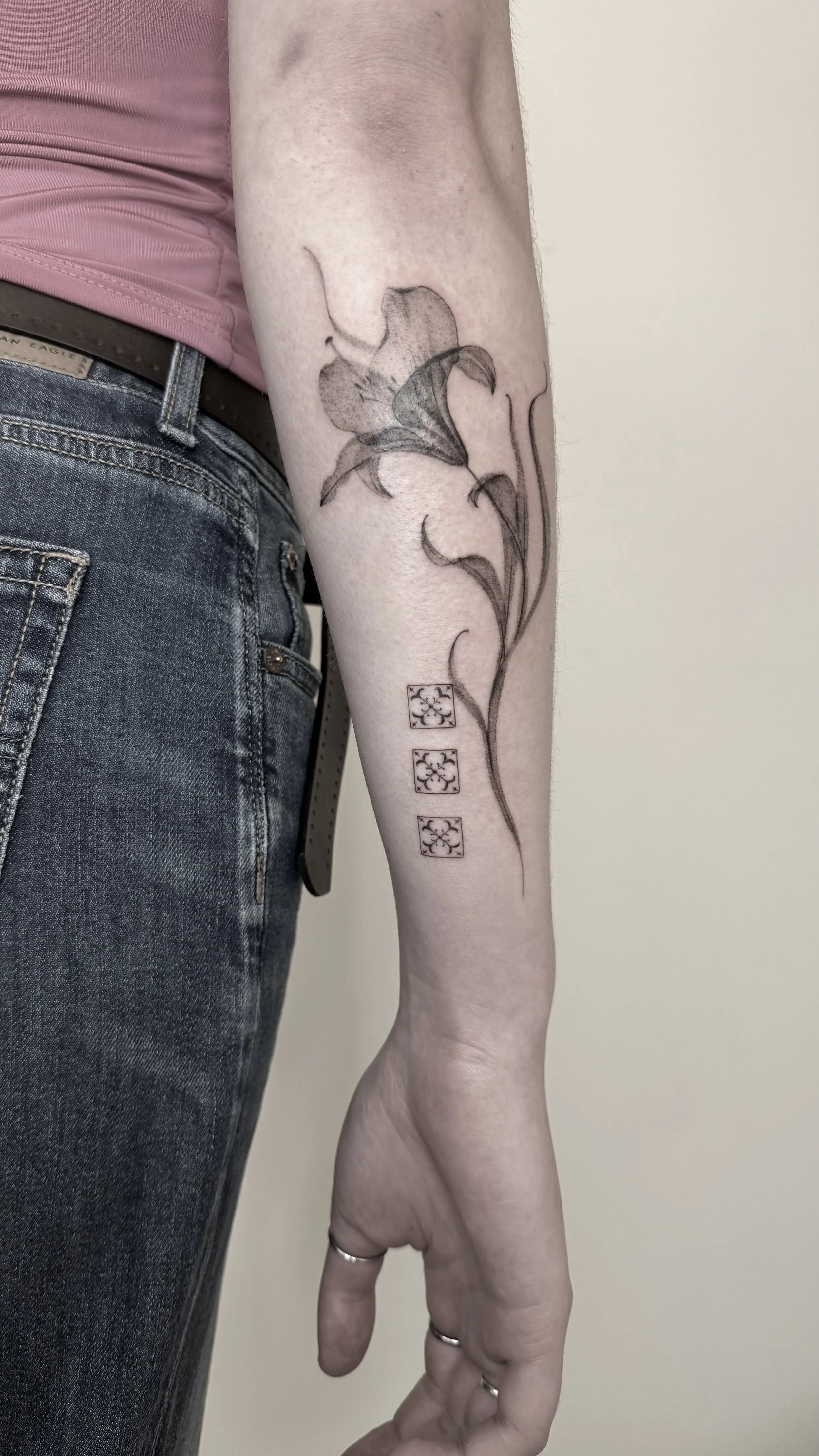 A tattoo of a lily flower with three small square designs underneath is on a person's forearm.