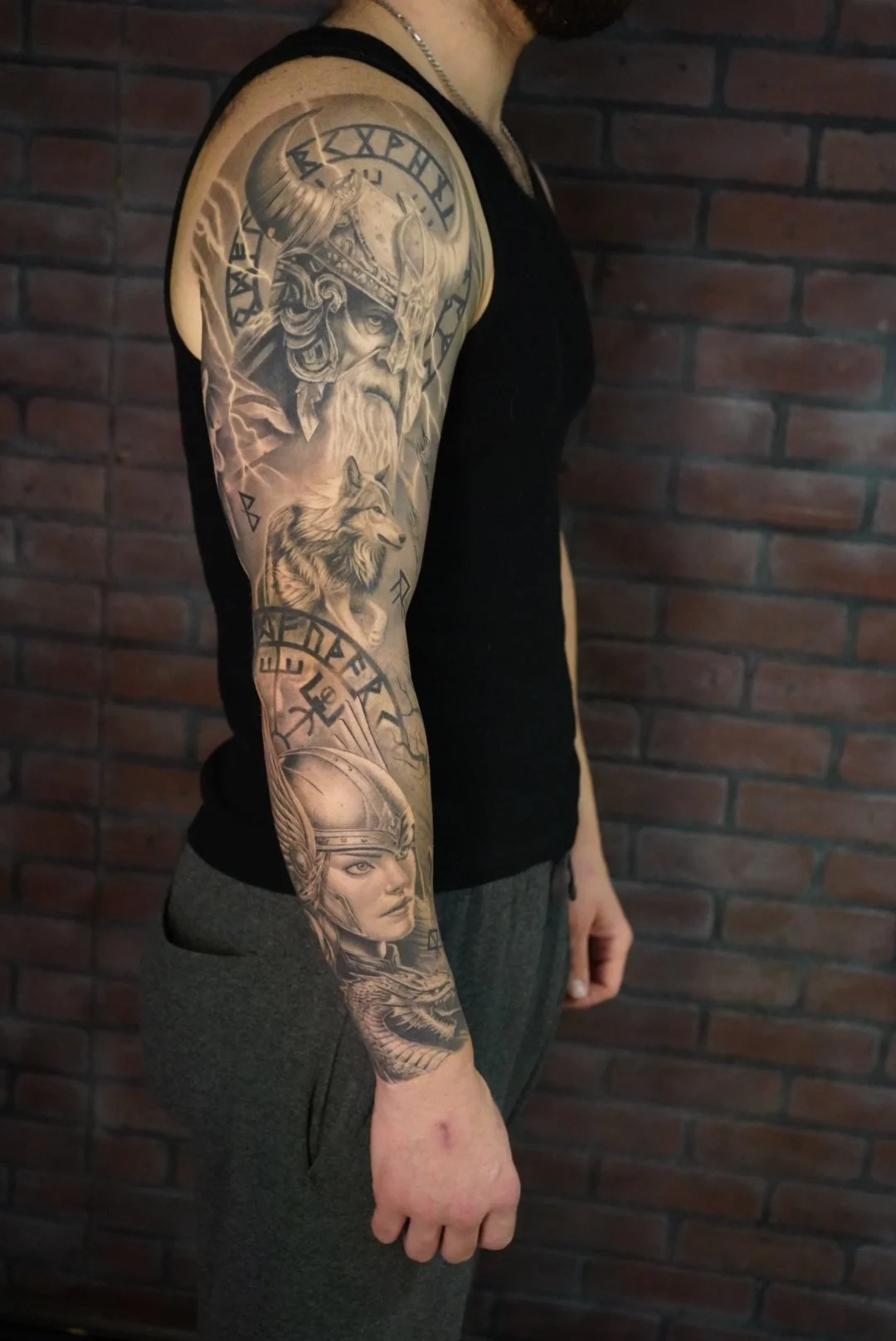 Tattoo sleeve on a man's arm featuring a Viking warrior with a horned helmet, a wolf, a woman with a helmet, and runic symbols, with a brick wall background.