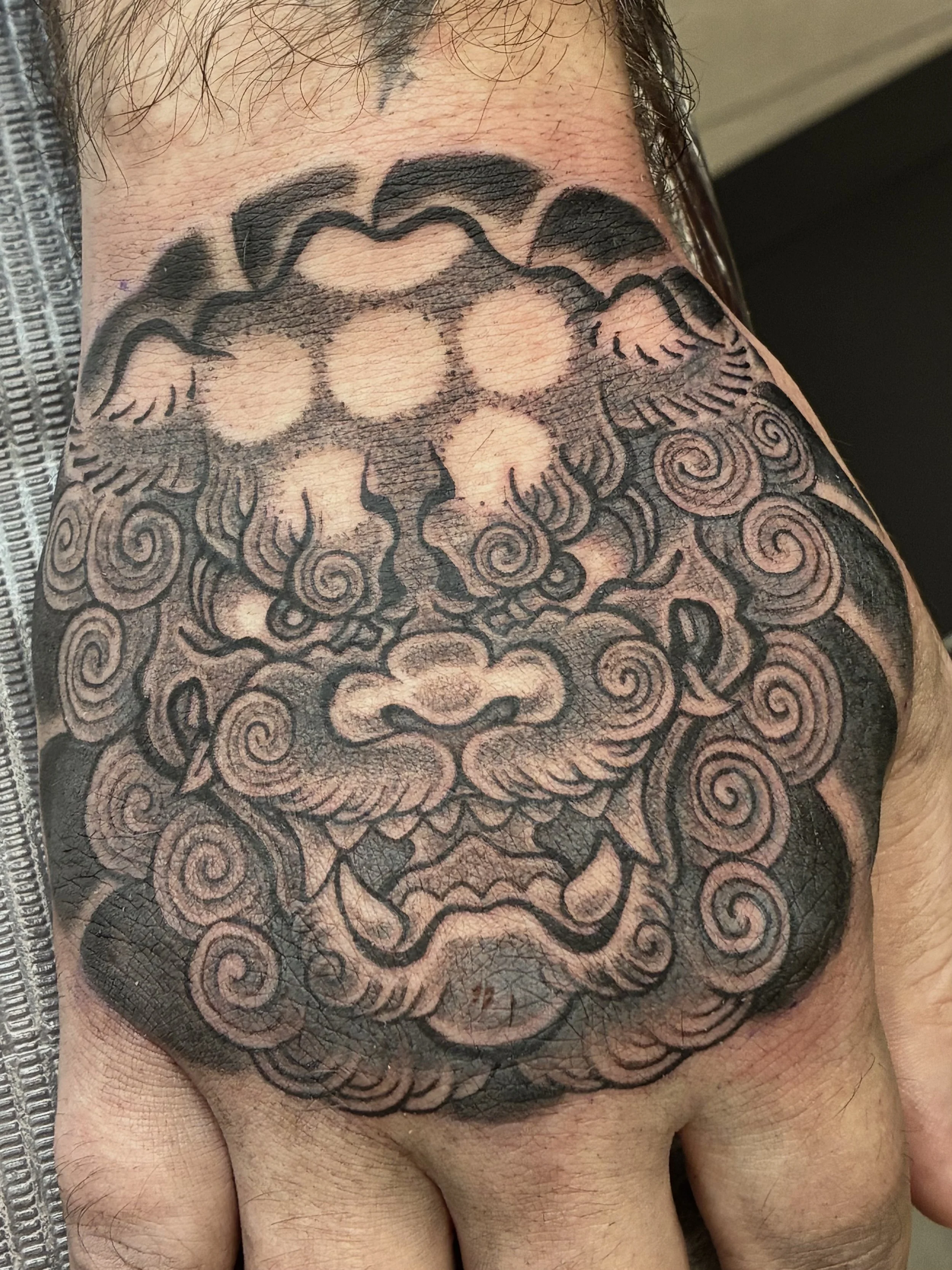 Close-up of a person's shoulder with a large tattoo featuring a traditional style design of a dragon's face with intricate patterns and swirling details.