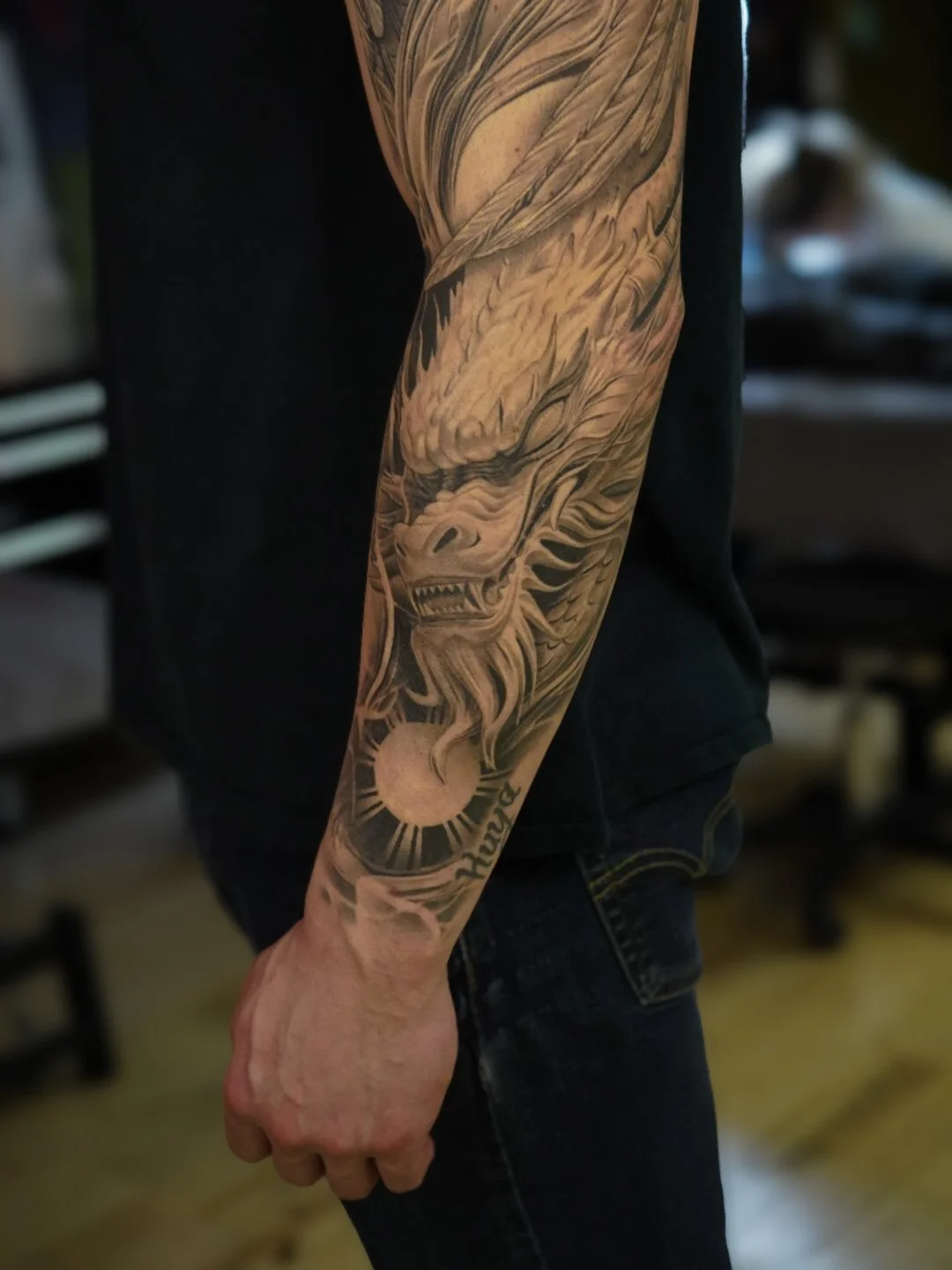 Close-up of a person's arm with a large, detailed black and gray tattoo of a dragon's head, clouds, a sun, and the word 'truth'.