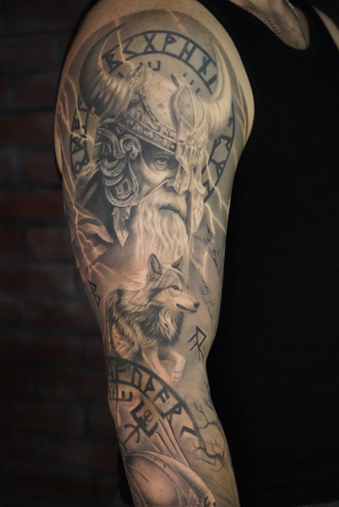 Tattoo of a Viking warrior with a horned helmet, beard, and stern expression, accompanied by a wolf, and runic symbols.