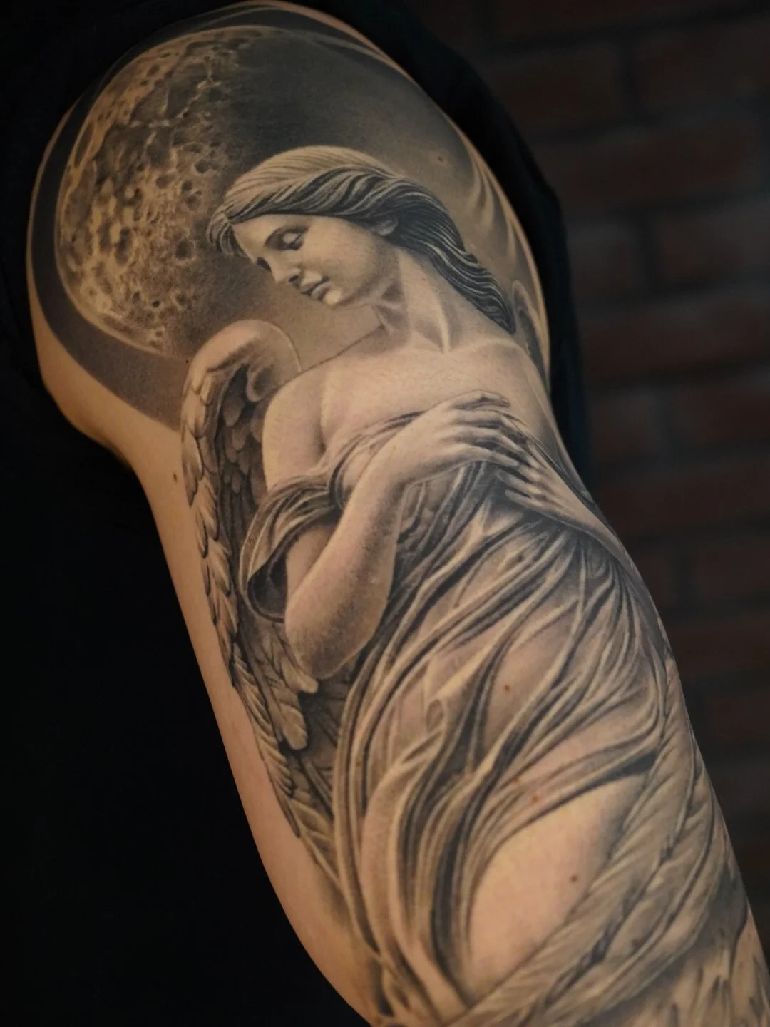 Black and gray tattoo of a woman with angel wings holding an orb, with a moon or celestial body in the background.