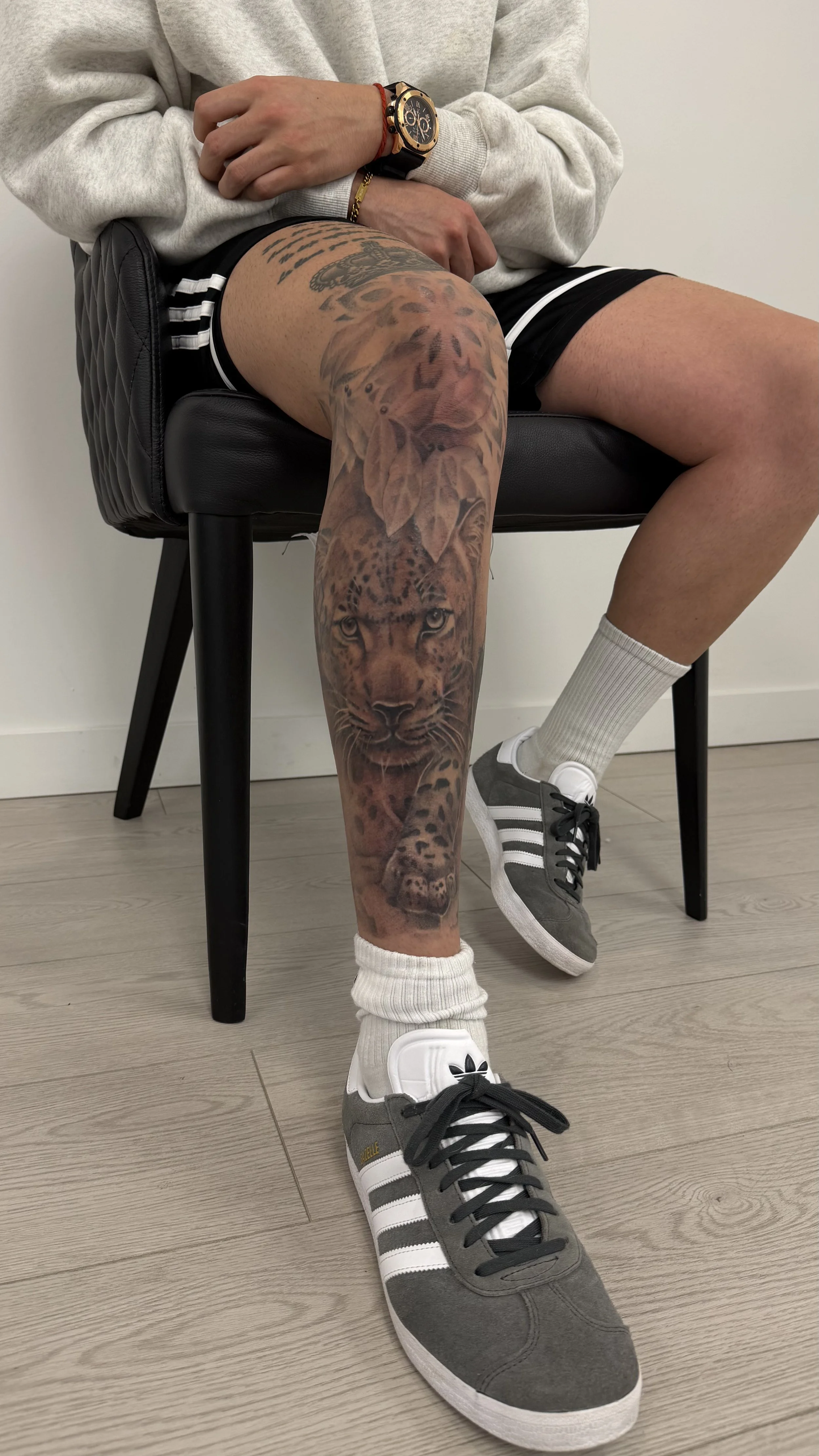 Person sitting on a black stool, displaying a large tattoo of a lion on their left leg, wearing sneakers, white socks, a gray hoodie, black shorts, and a watch.