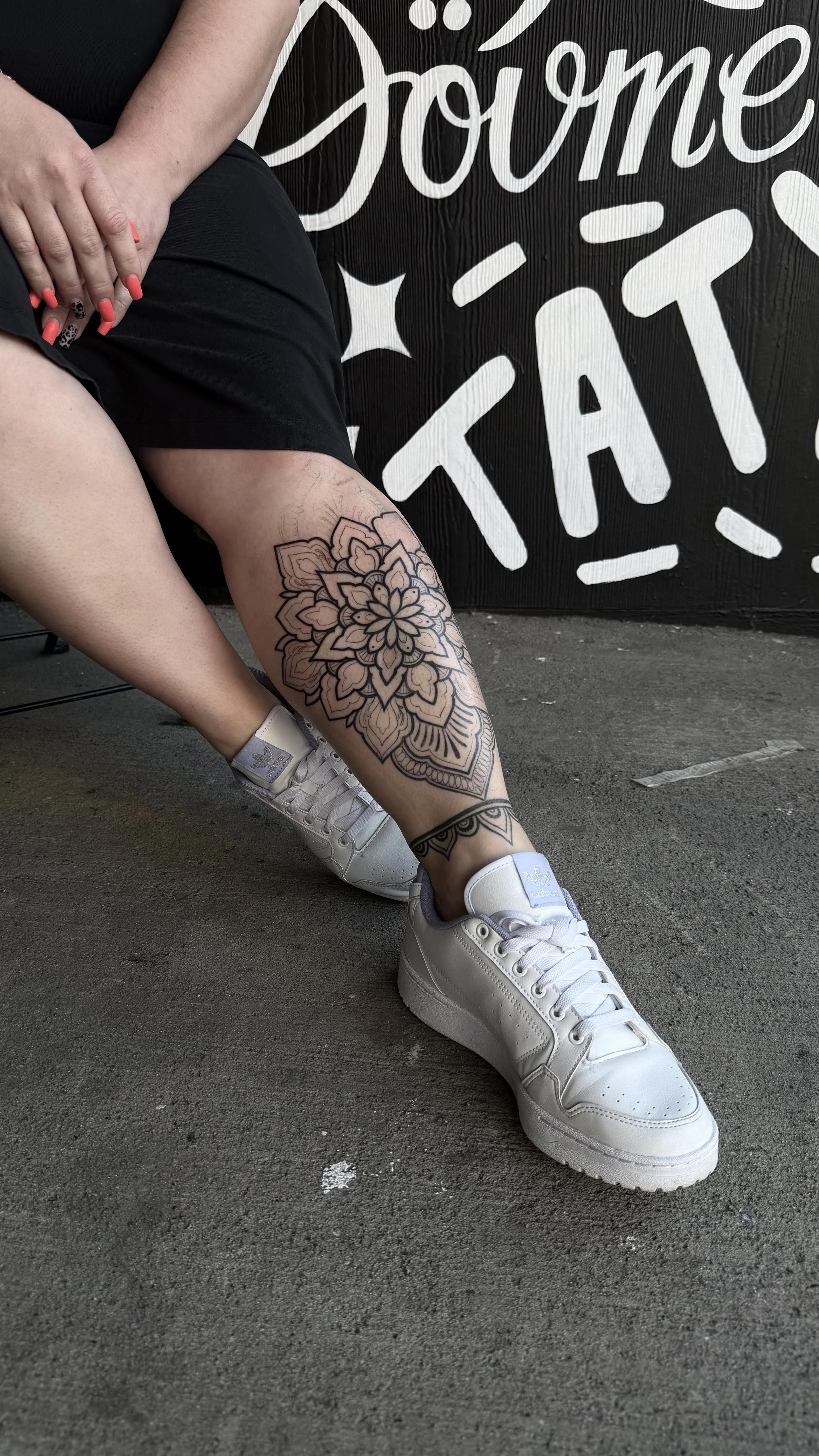 A person with neon pink nail polish sitting on the ground, showing a large floral tattoo on their leg, wearing white sneakers and black shorts, with a black background that has white lettering.