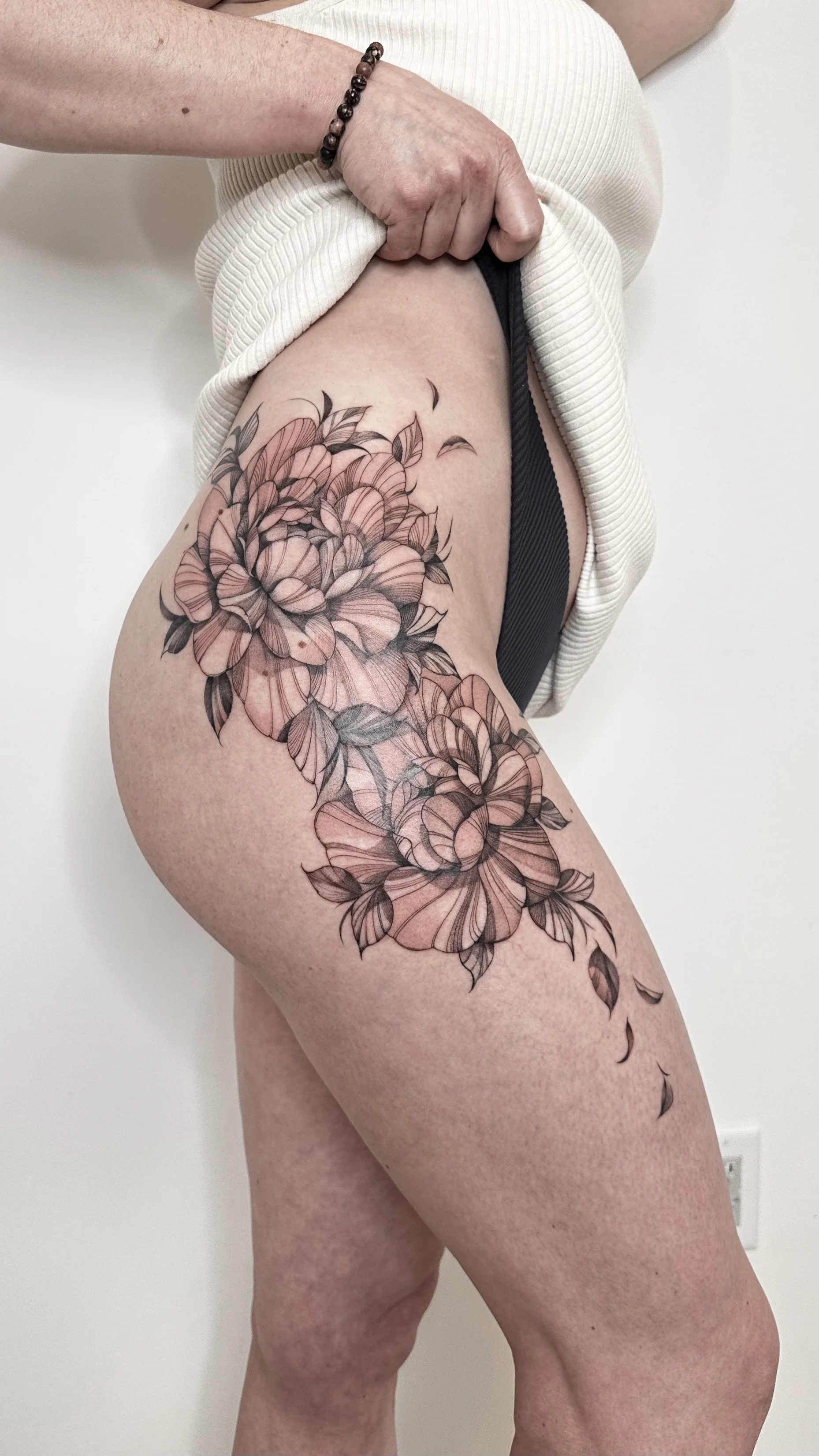 A person with a floral tattoo on their thigh, pulling up their cream-colored top to reveal the tattoo, wearing black underwear and a beaded bracelet.