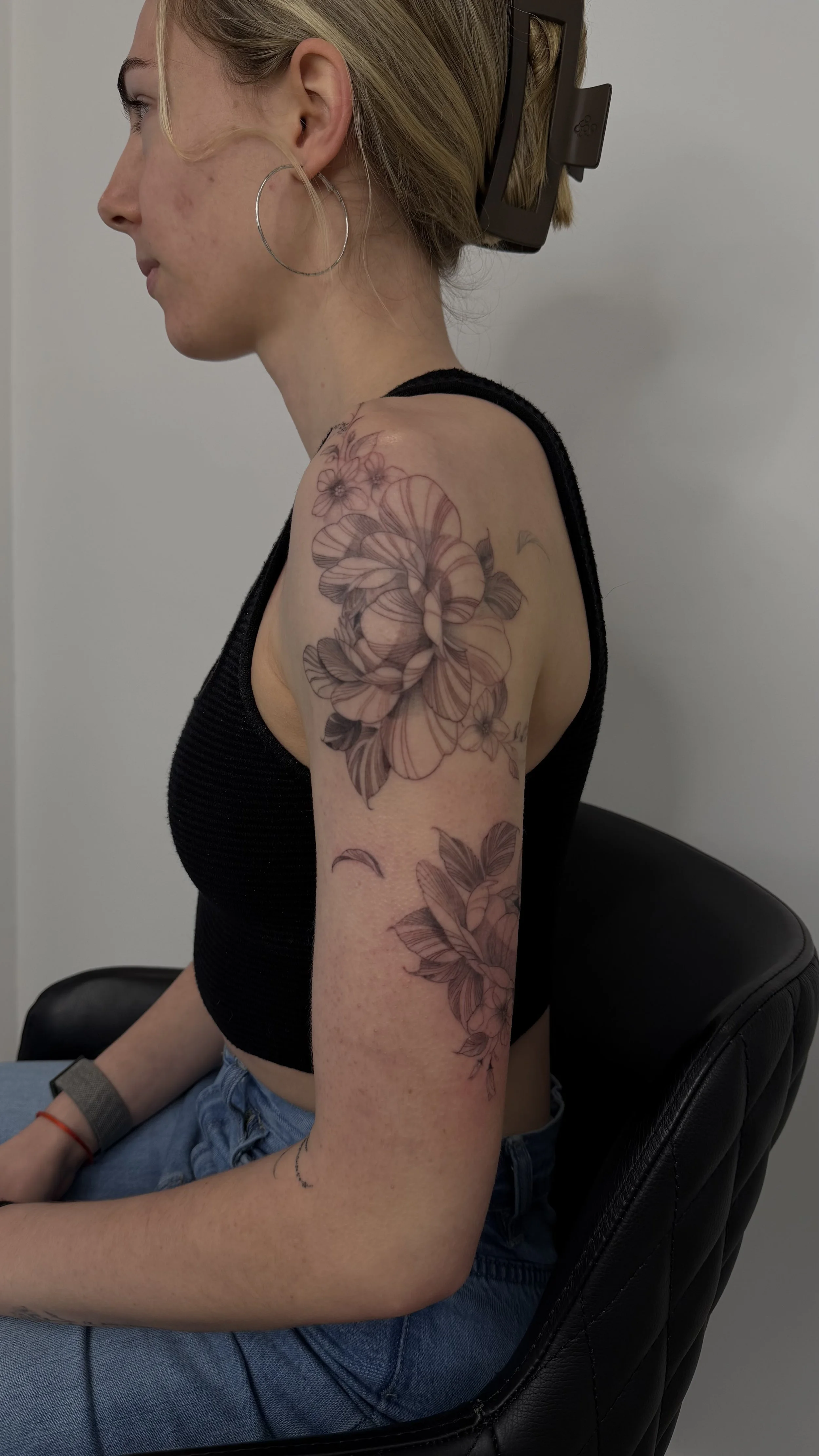 Side view of a woman sitting in a black chair, wearing a black sleeveless top, blue jeans, earrings, and a hair clip, with a floral tattoo on her arm.