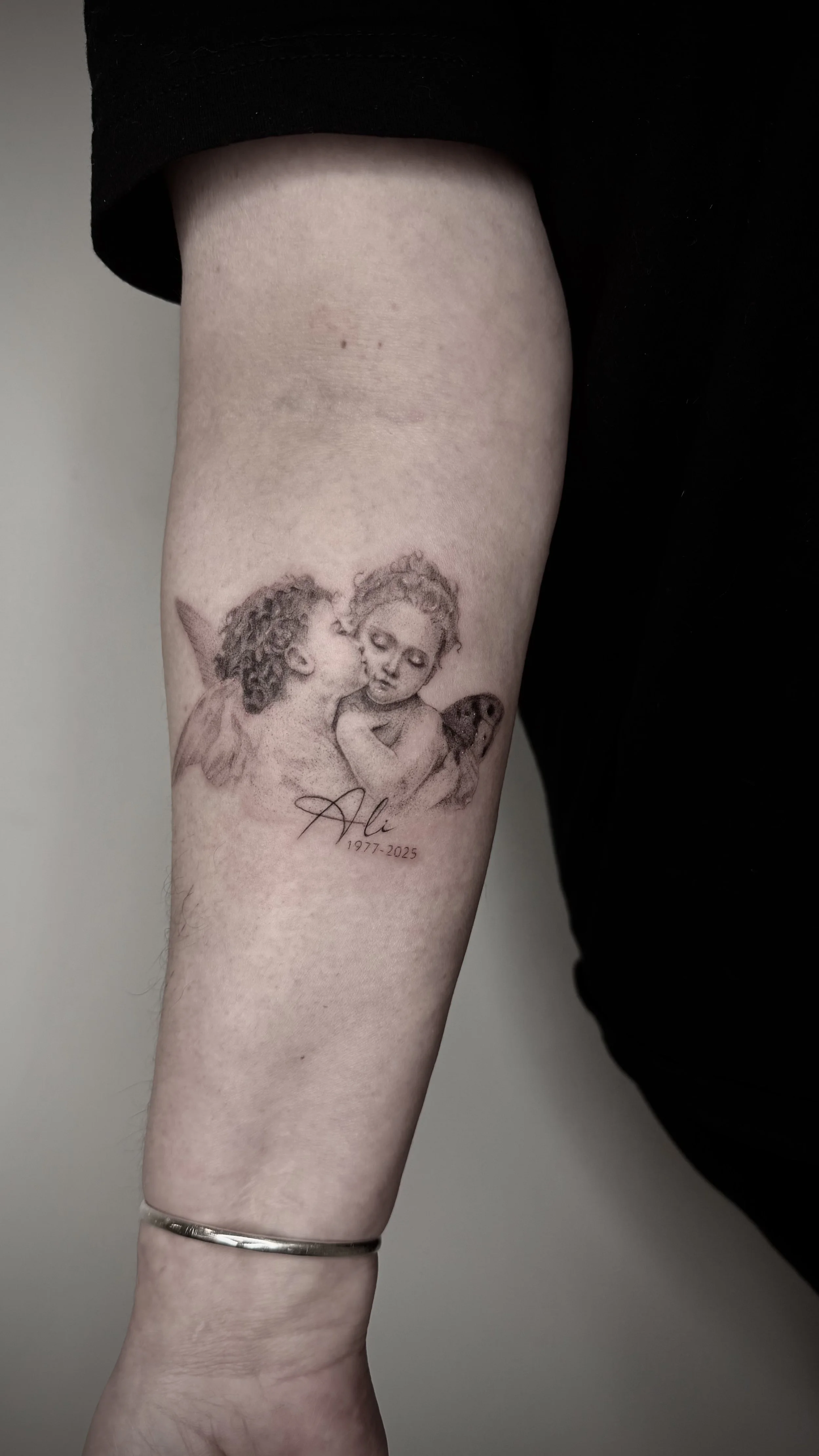 Tattoo of a boy and girl with angel wings, kissing, with the name 'Ala' and dates 1977-2025 underneath.