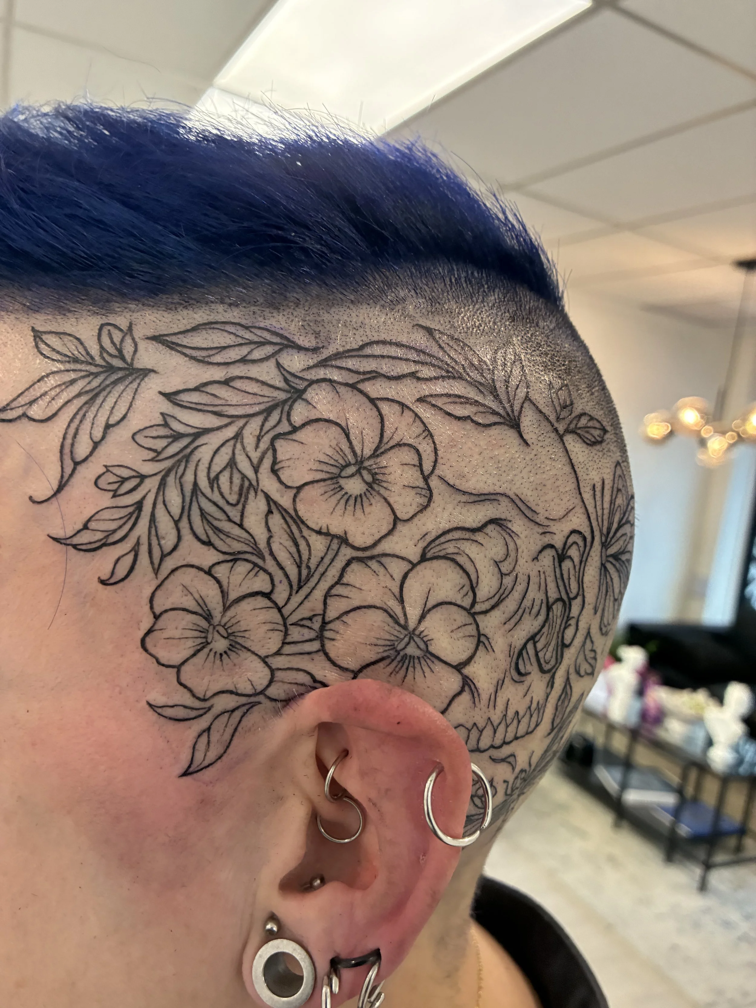 Close-up of a person's head showing short, dark hair with a floral tattoo design on the scalp.