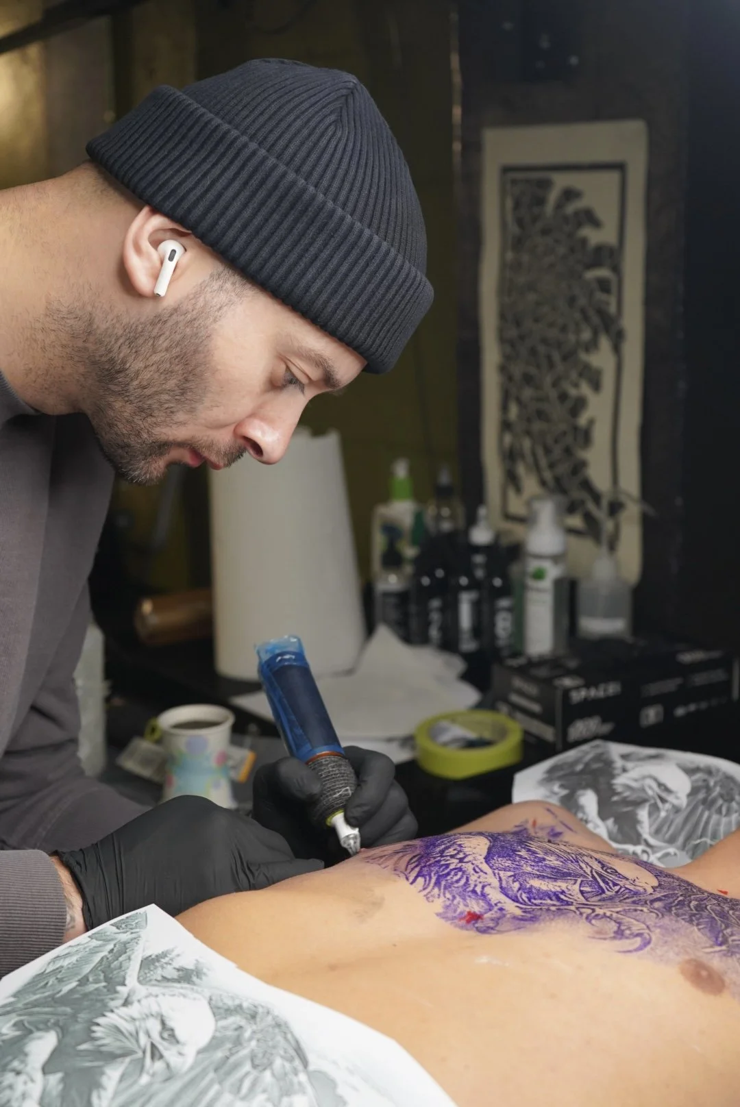 Tattoo artist wearing a beanie, black gloves, and wireless earbuds, tattooing a detailed design on a person's back in an art studio.