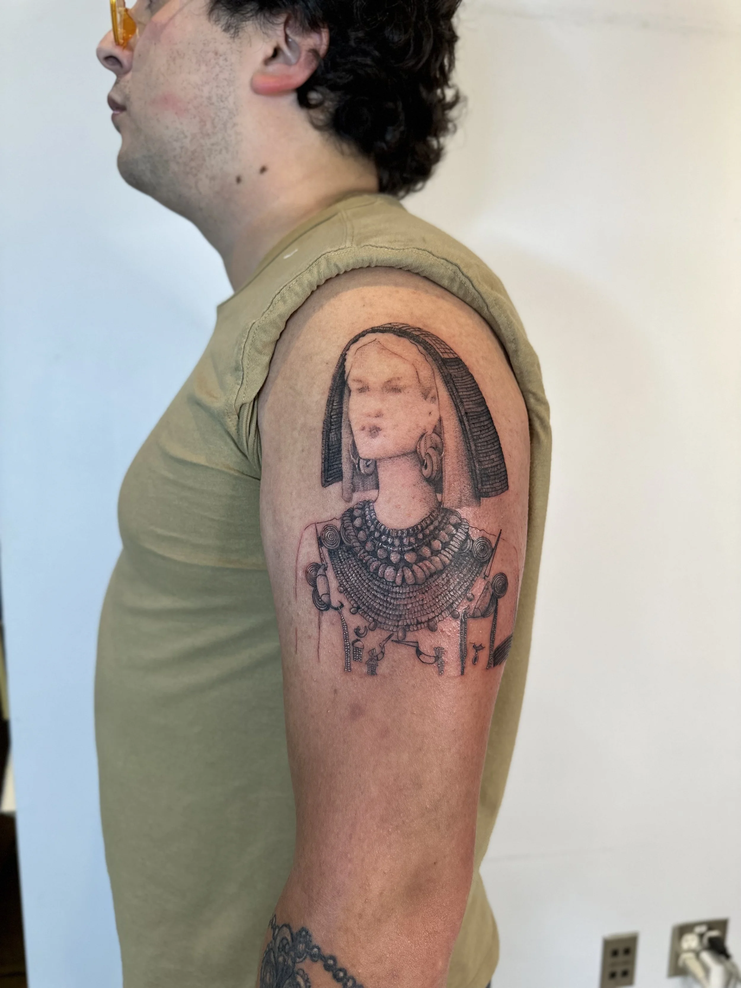 A man with dark, curly hair and a fair complexion shows off a tattoo on his upper arm of an Egyptian queen in profile, wearing a headdress and ornate jewelry.