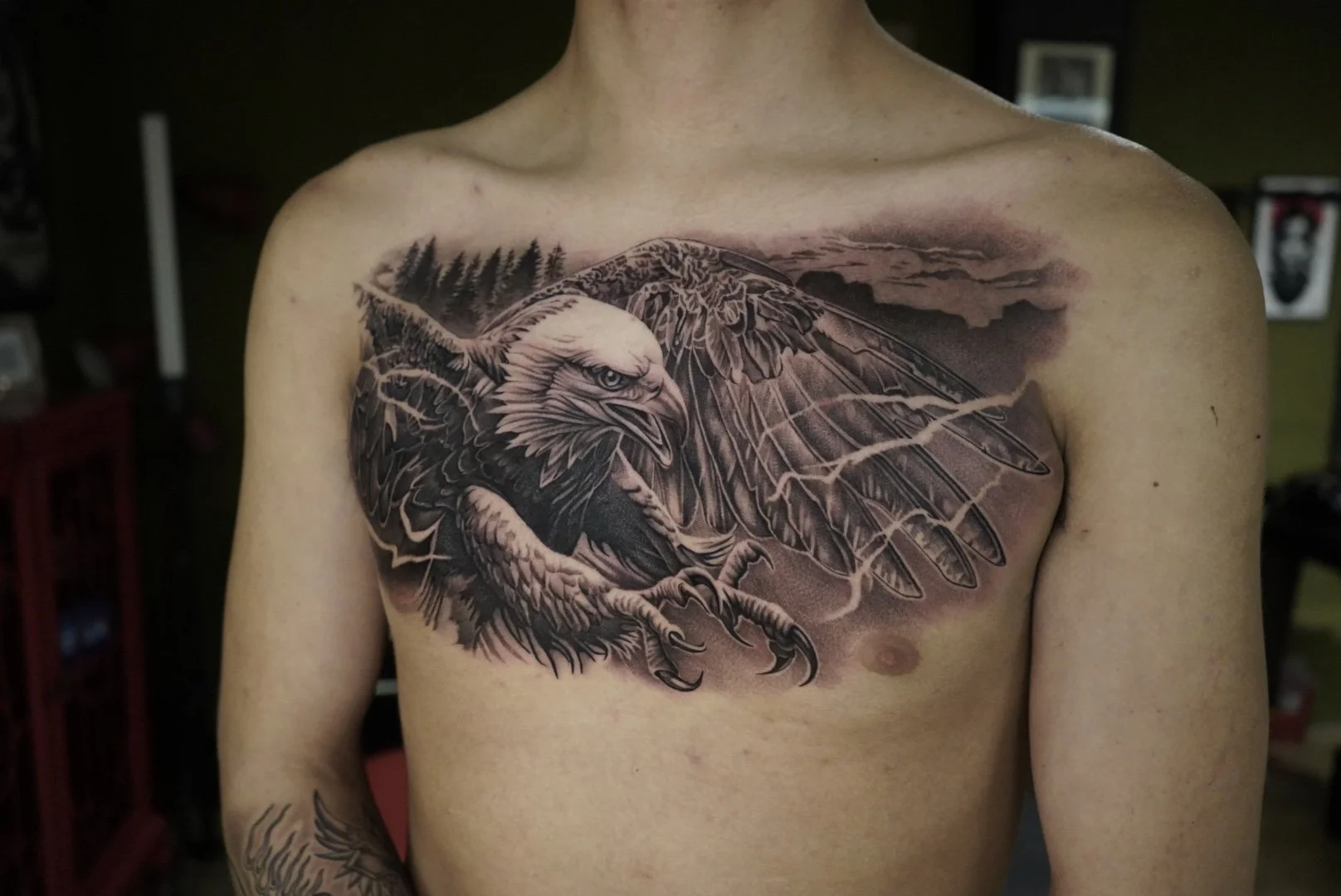 Black and gray realistic tattoo of an eagle with its wings spread, depicted on a person's chest. The background includes clouds and mountains.