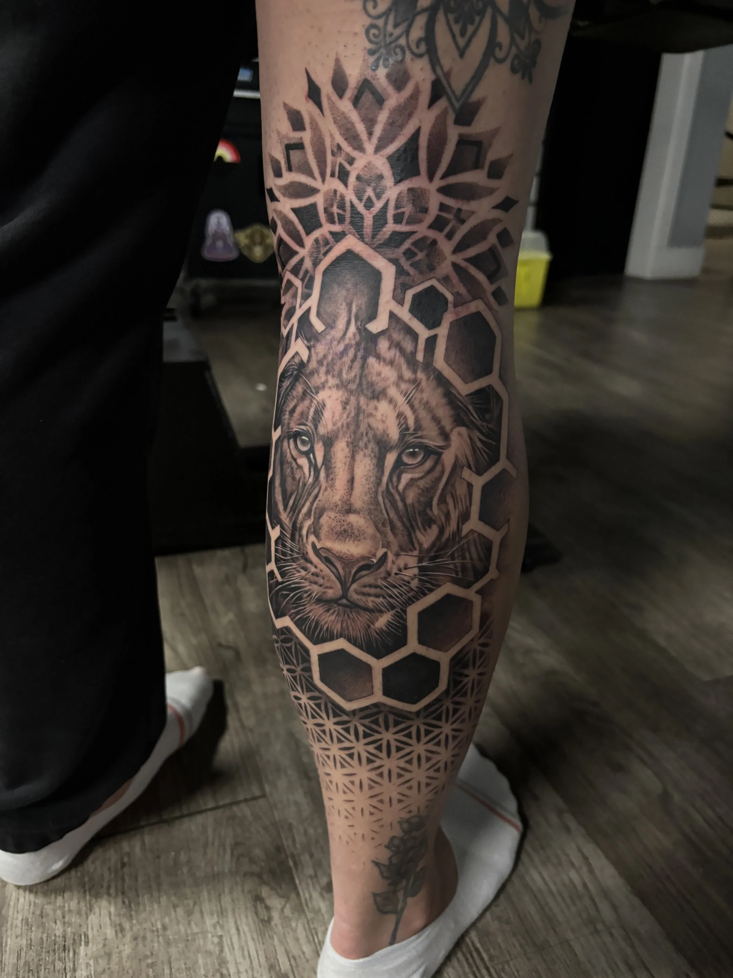 Tattoo of a lion's face with detailed shading, top geometric honeycomb pattern, and lower floral design on a person's lower leg.