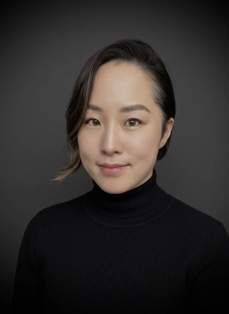 A woman with dark, shoulder-length hair styled to one side, wearing a black turtleneck, posing against a dark gray background.