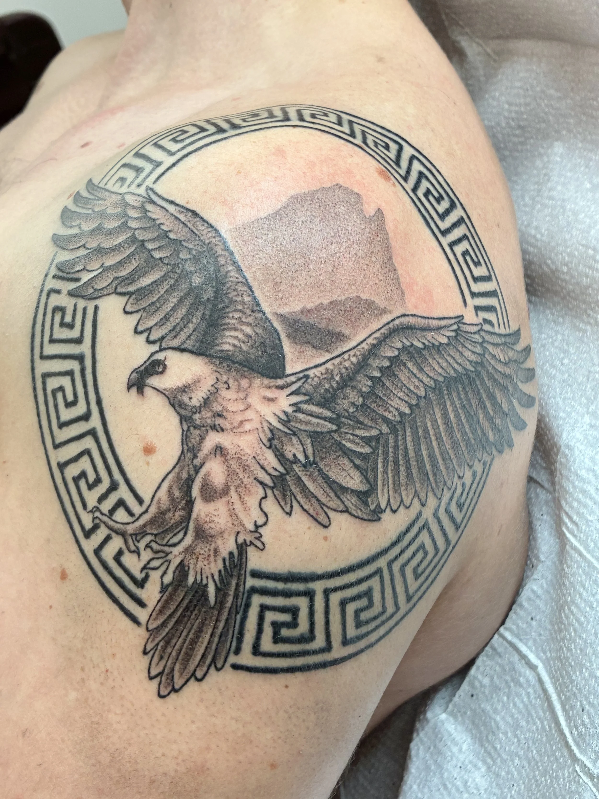 Tattoo of an eagle inside a circular Greek key pattern on a person's shoulder with a mountain landscape in the background