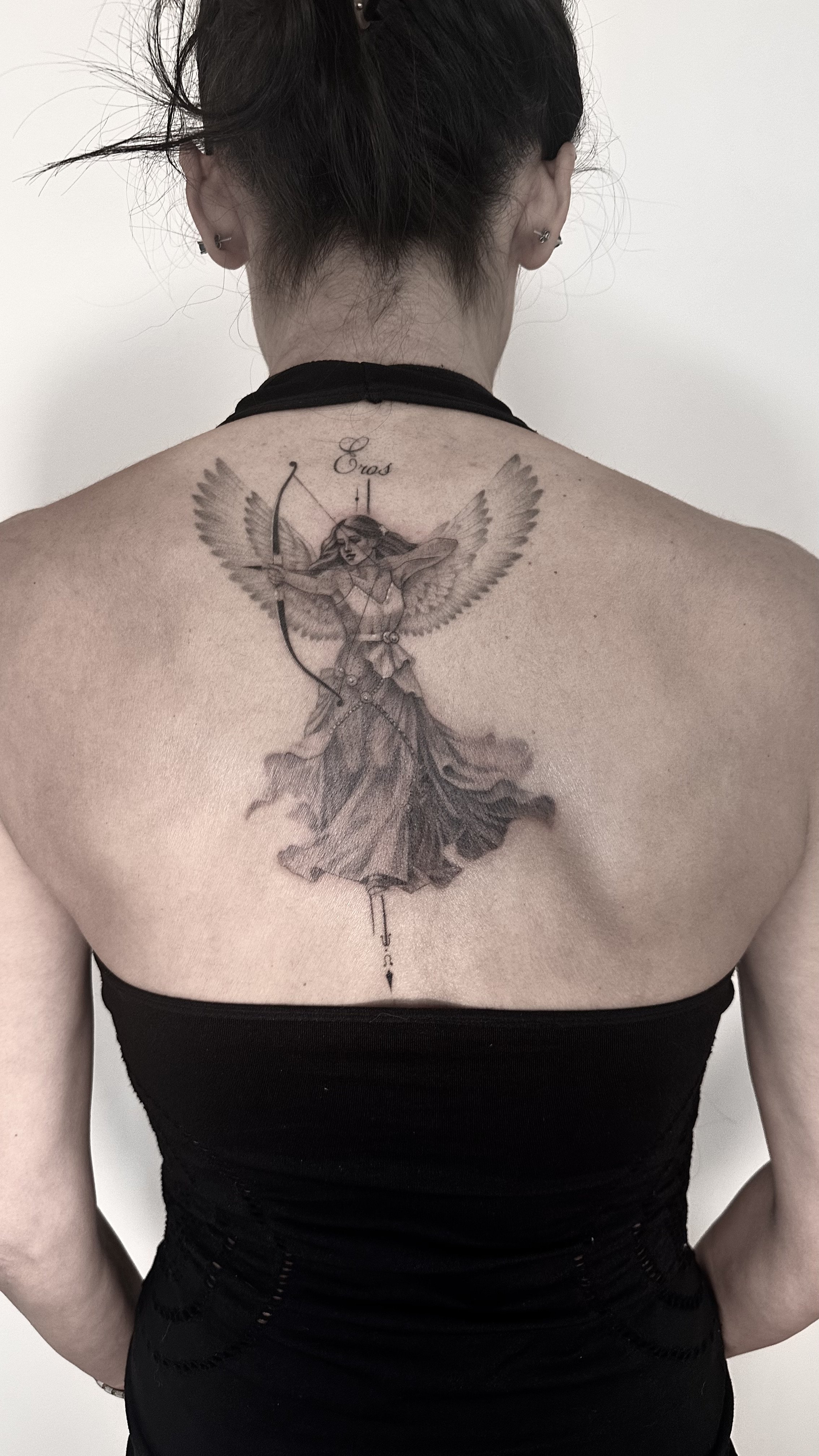 Back view of a person with curly hair and earrings, wearing a black top, revealing a detailed tattoo of a winged woman holding a bow on their upper back.