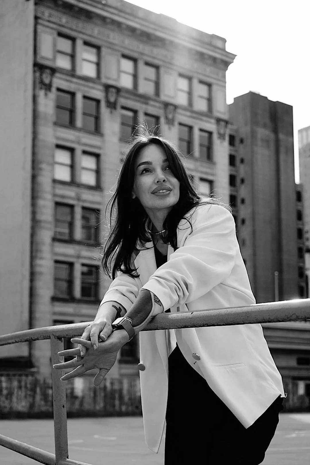 Black and white photo of a woman with dark hair leaning on a railing outdoors, with tall city buildings in the background.