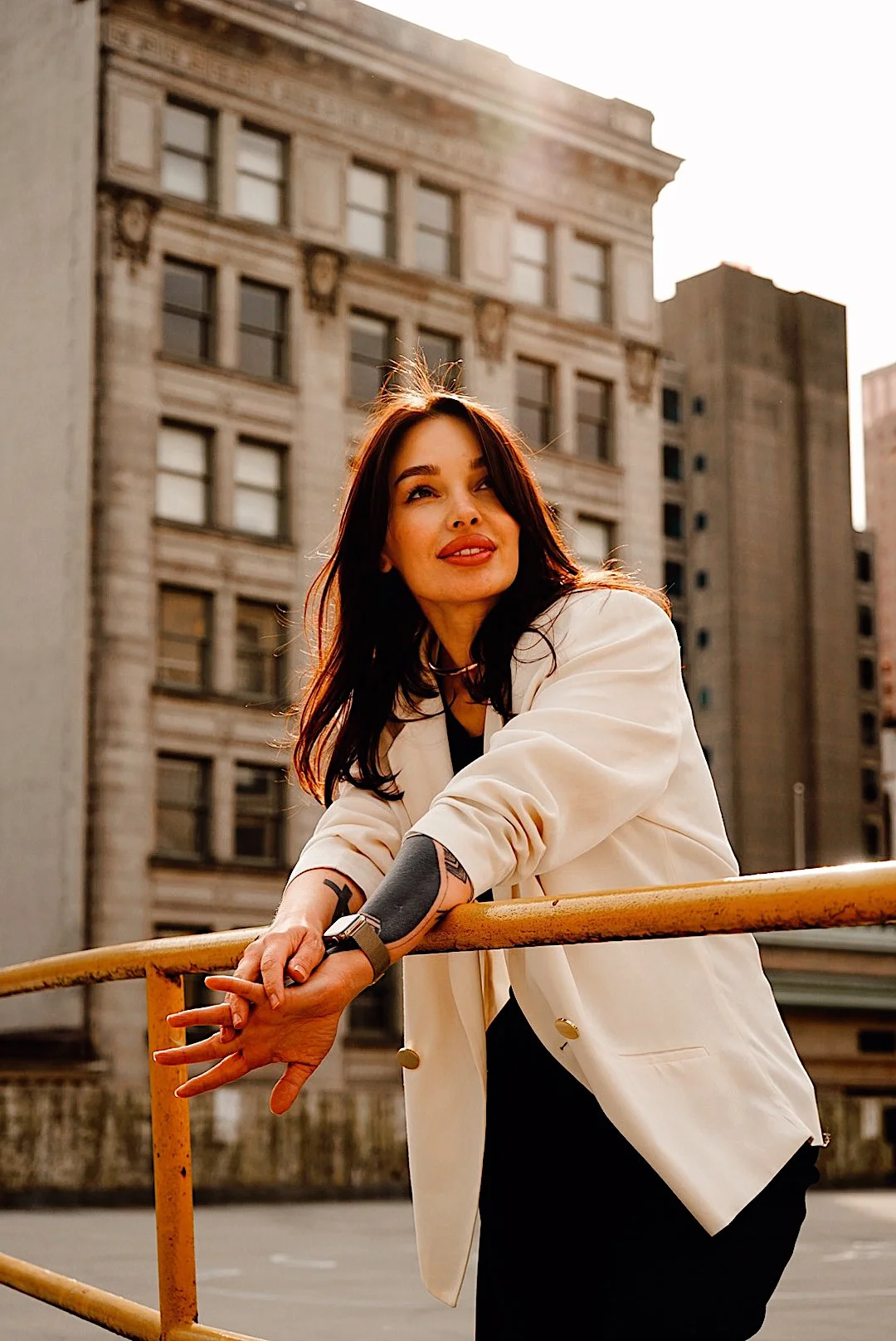 A woman with dark hair leaning on a yellow railing in an urban setting, wearing a white blazer and black shirt, with sunlight behind her and historic building in the background.