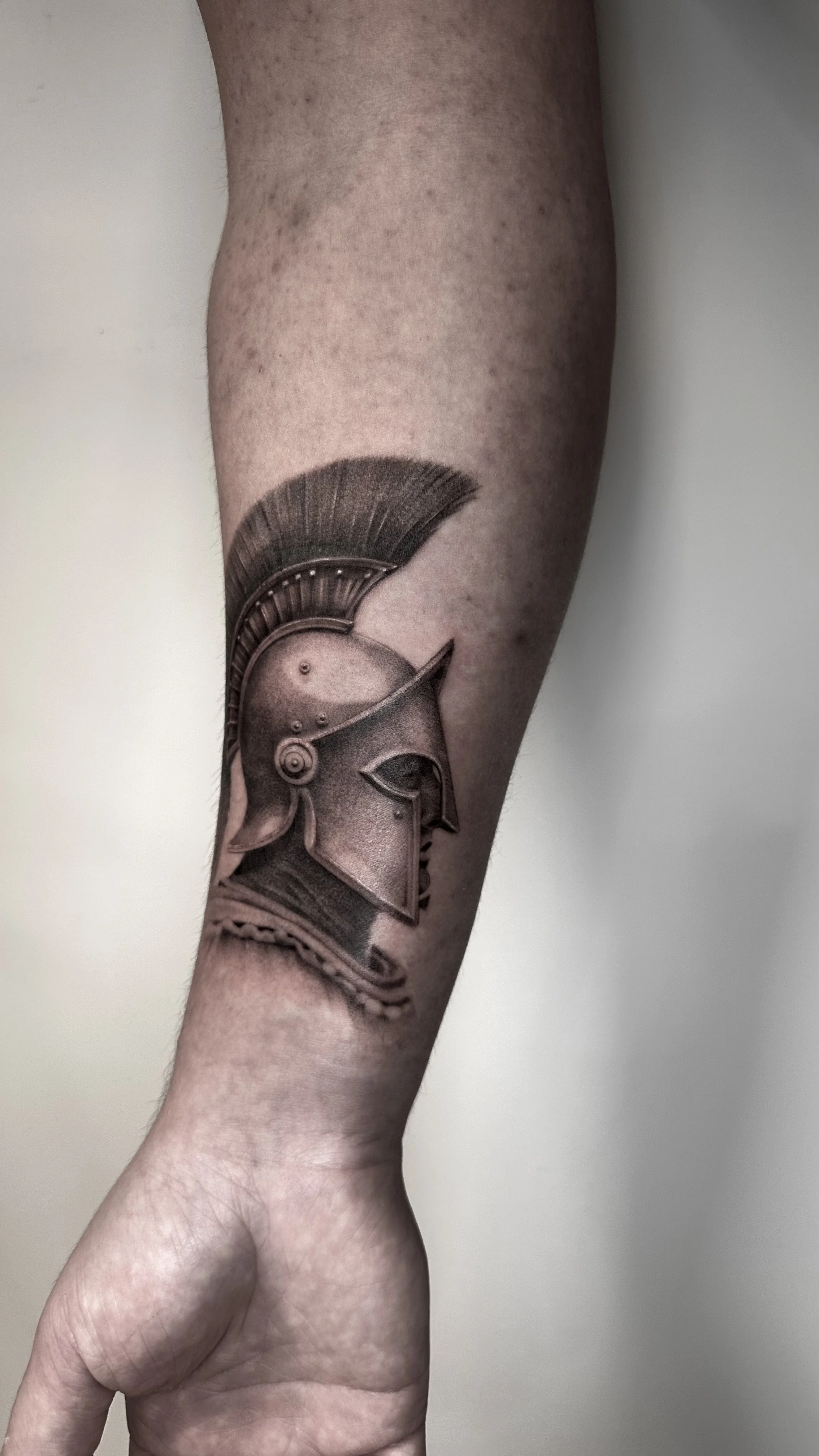 Tattoo of a classical Greek or Roman soldier's helmet on a person's forearm.